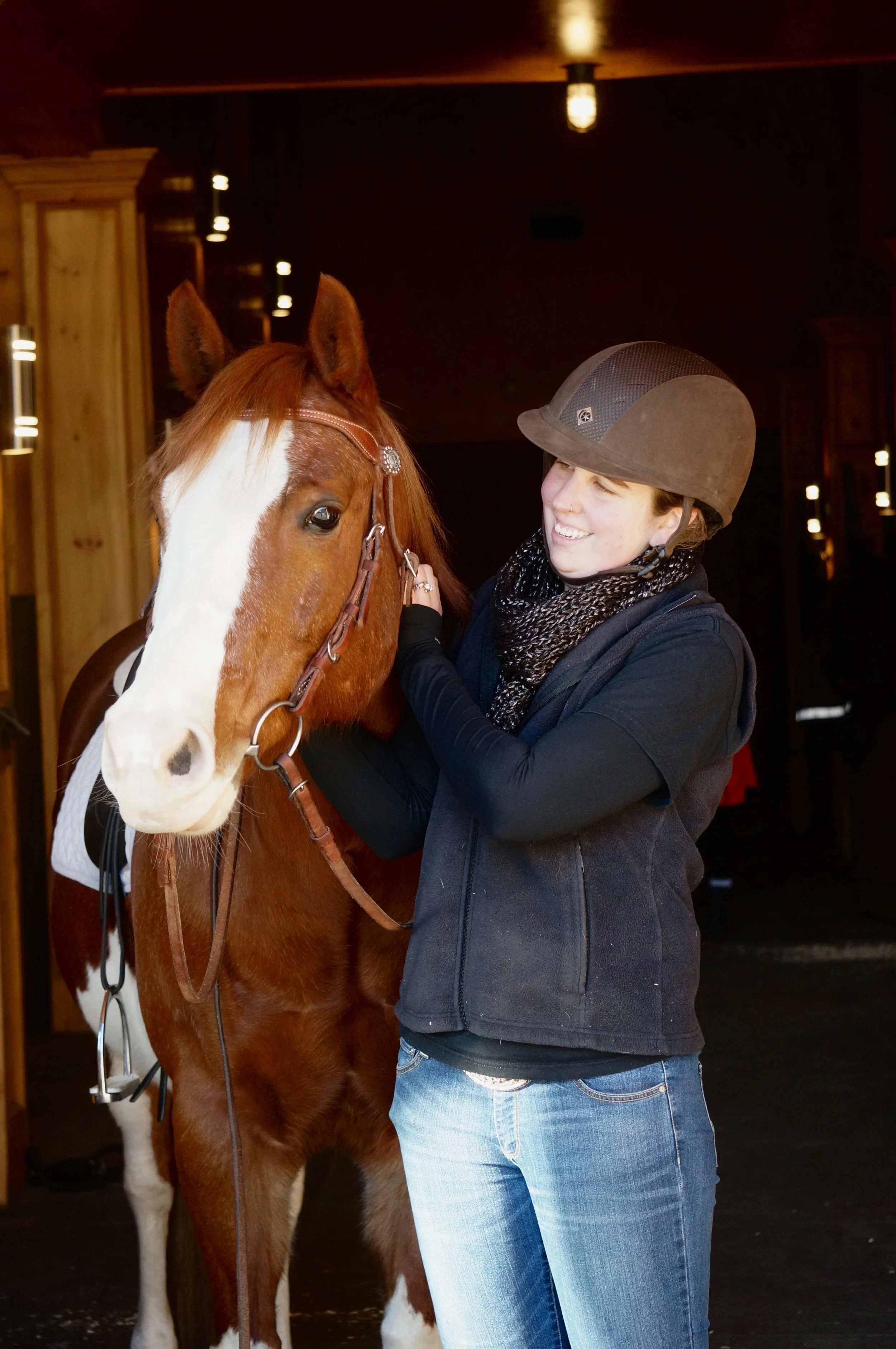 A young woman wearing a riding helmet and a scarf smiles as she stands next to and touches the face of a brown horse with a white blaze.