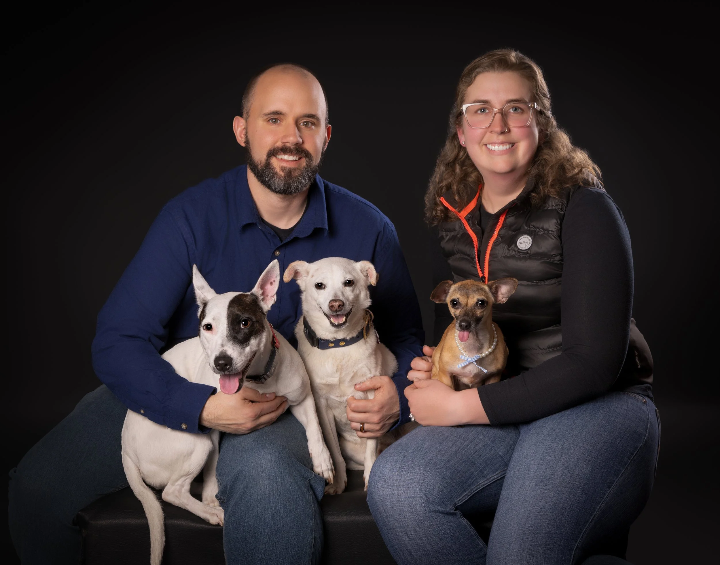 A man and woman sitting on a dark background, each holding two dogs. The man has a beard and wears a navy blue shirt, and the woman has curly hair, glasses, and a black vest. They are smiling at the camera.