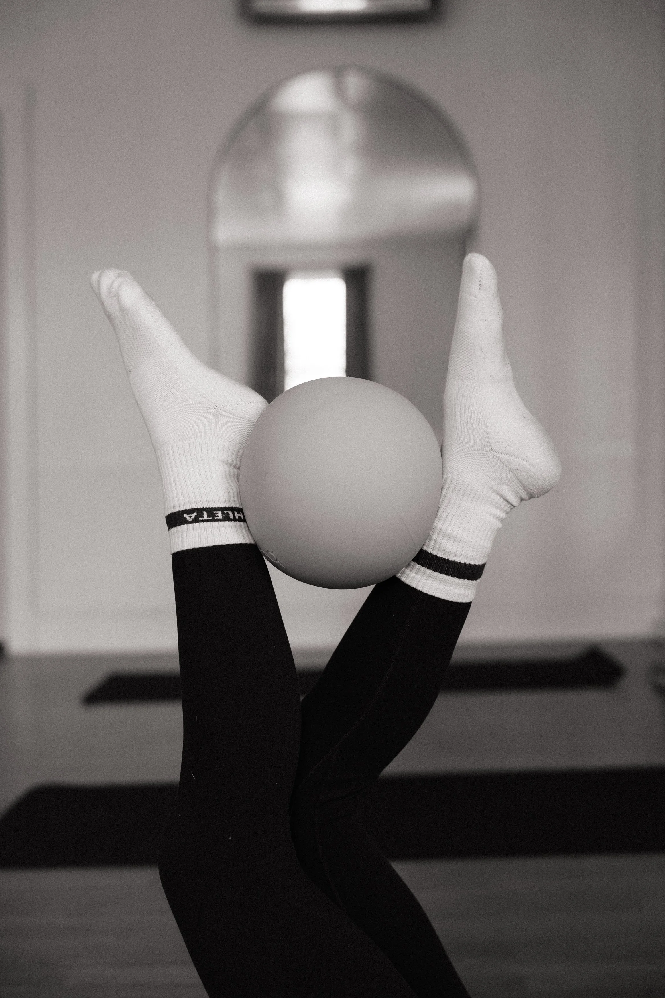 A person in black athletic clothing, wearing white gloves, is doing a yoga pose with feet pointing upward and holding a white sphere between their legs. The background features a mirror reflecting a blurred window.