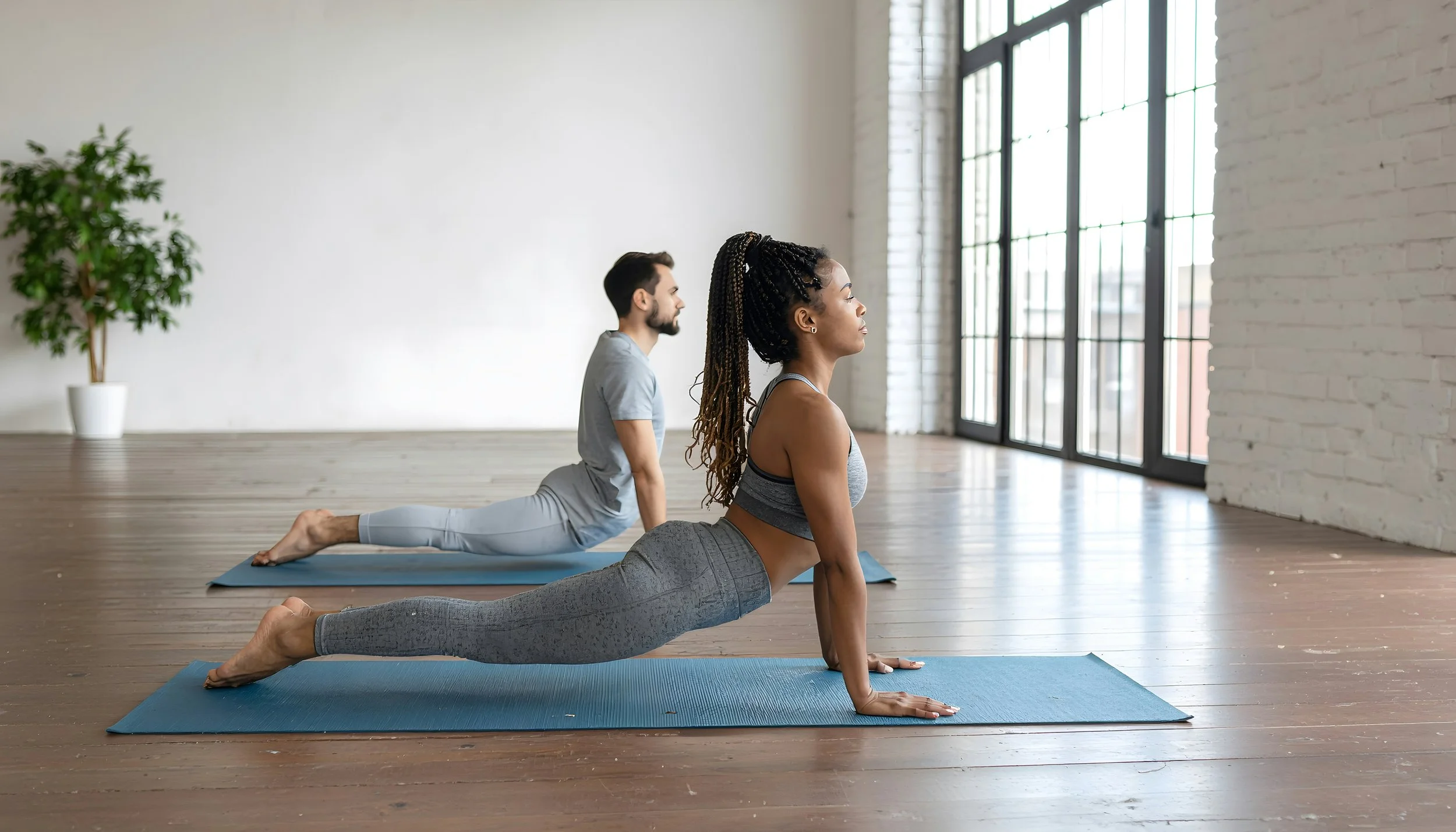 A woman and a man practicing yoga in a spacious studio with large windows and wooden floors, performing cobra pose on yoga mats.