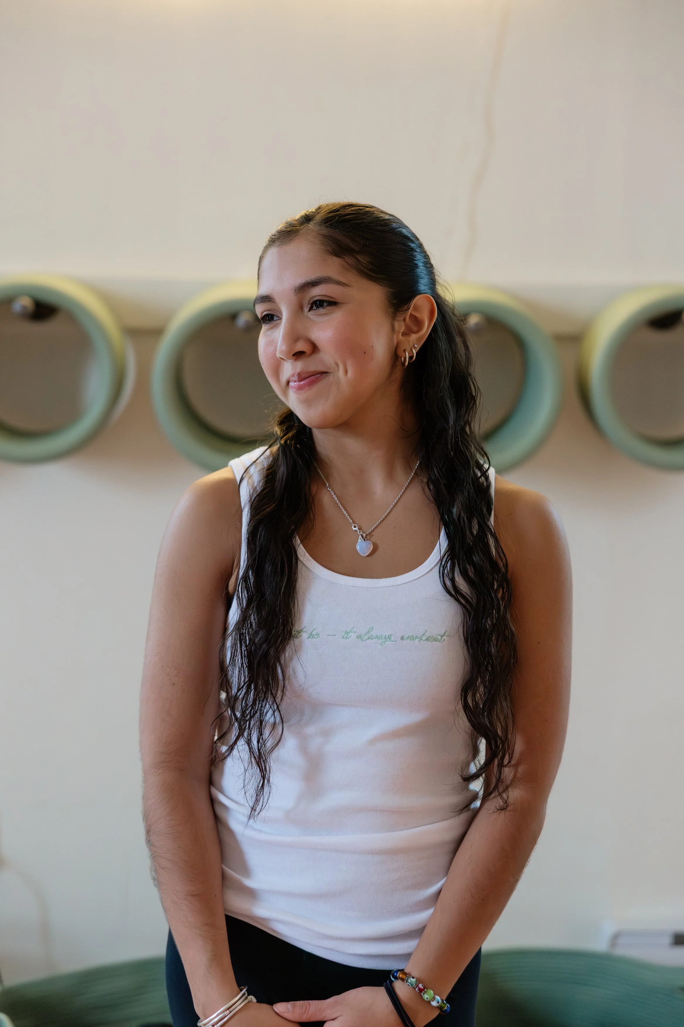 A young woman with long wavy hair, wearing a white tank top and multiple bracelets, looking to her right and smiling softly, standing in front of a wall with decorative circular objects.