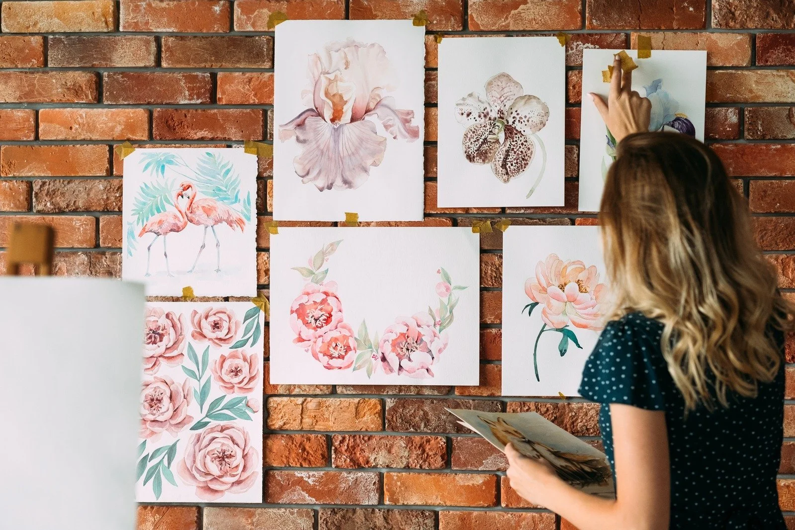 A woman with long wavy hair in a polka-dot shirt looks at watercolor paintings of flowers and animals taped to a brick wall, holding a booklet in her hands.