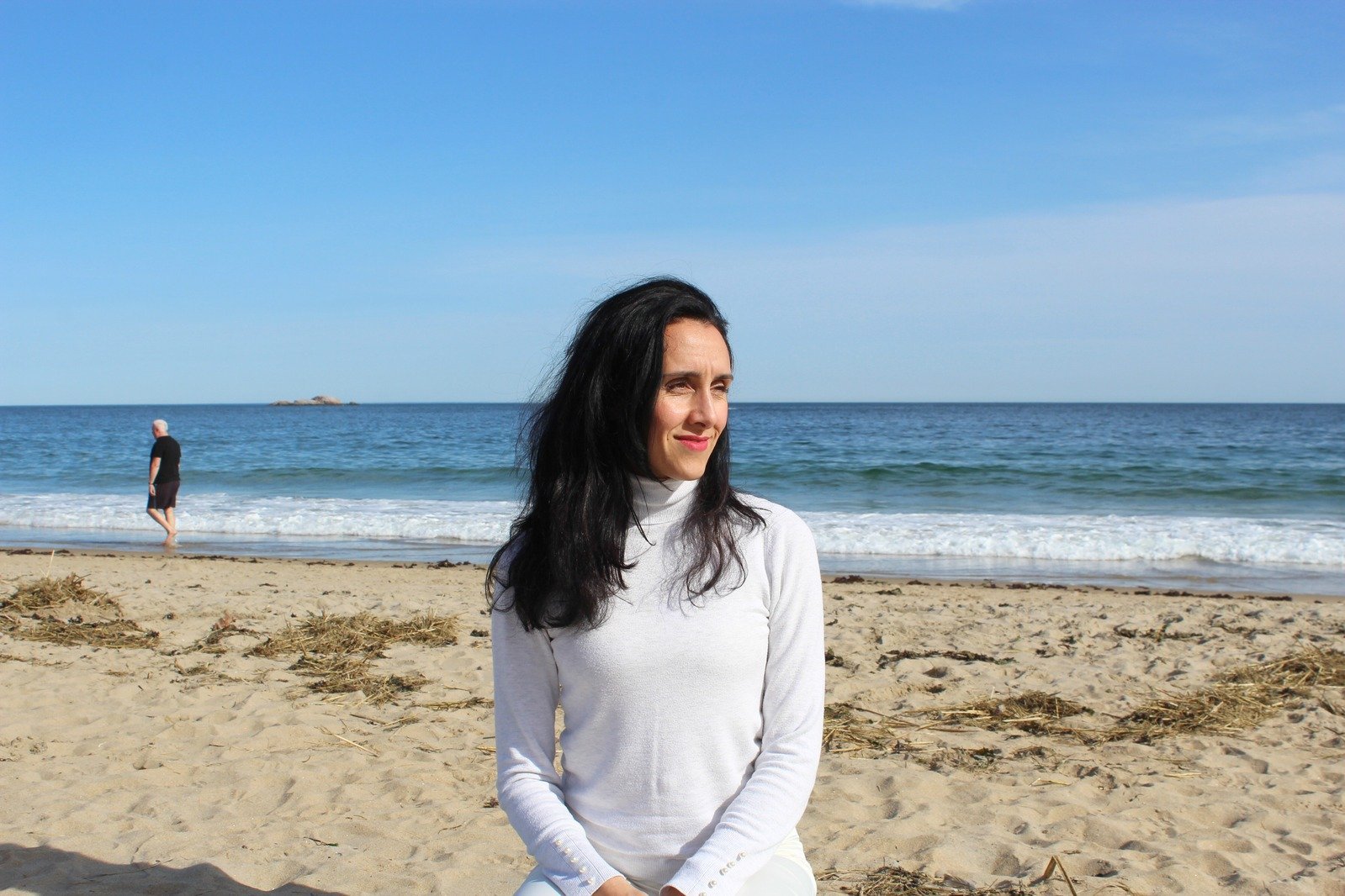 A woman with black hair wearing a white turtleneck sitting on the sandy beach with the ocean in the background, and a man walking near the water.