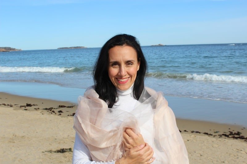 A woman with dark hair smiling at the camera on a beach, with the ocean and blue sky in the background, wearing a white top and a light pink shawl.