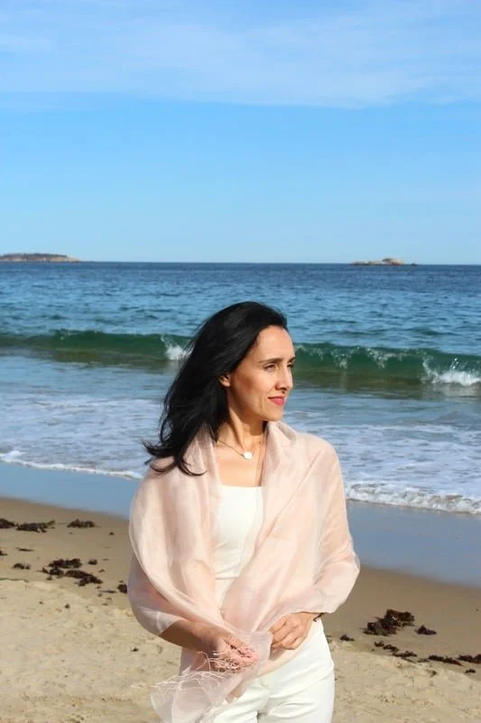 A woman with black hair stands on a sandy beach near the ocean, facing sideways with a thoughtful expression, wearing a light beige shawl over white clothing, with the sea and a clear blue sky in the background.