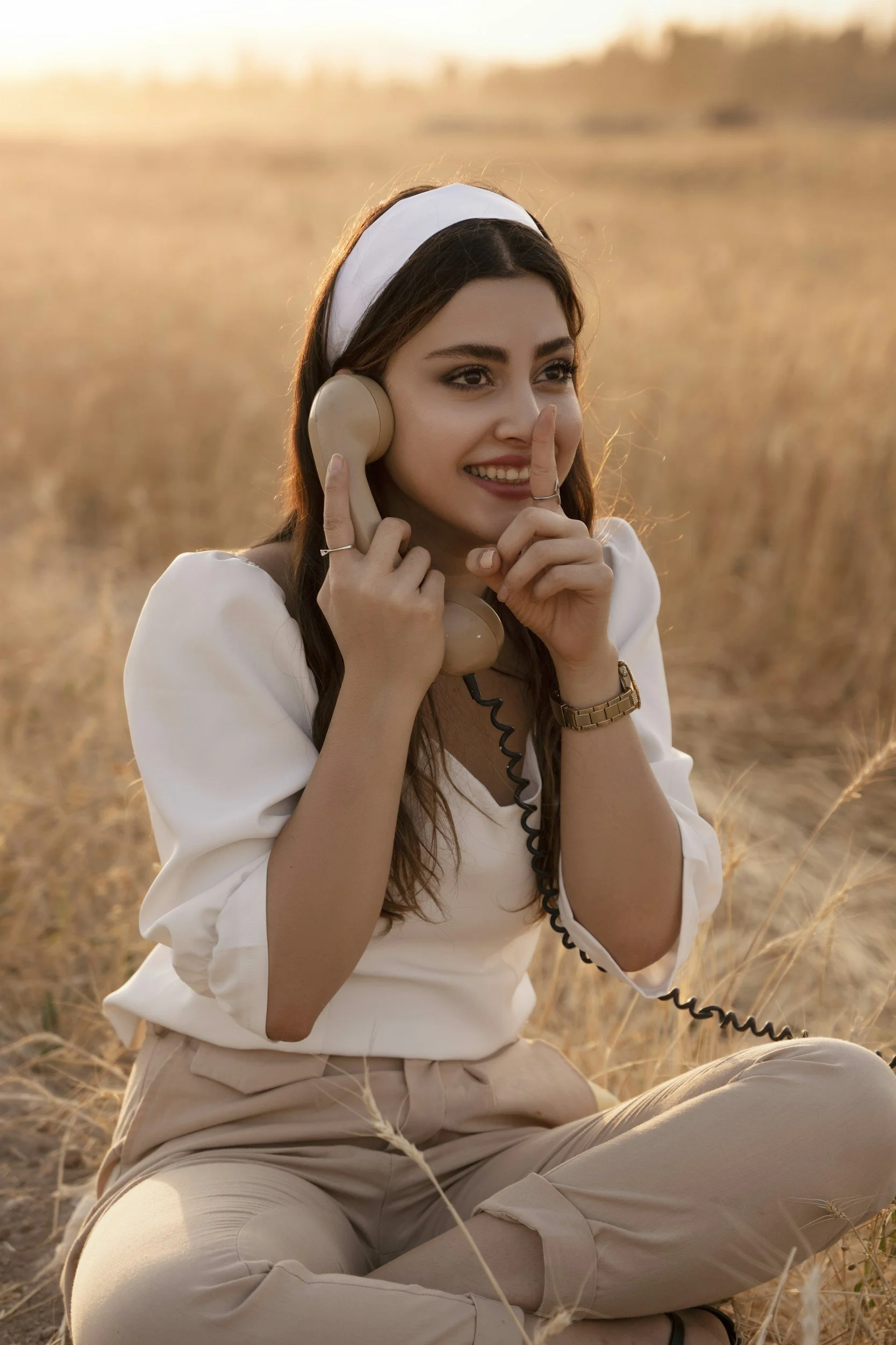 A young woman sitting in a field at sunset, holding a vintage telephone receiver to her ear, smiling and making a peace sign with her other hand.