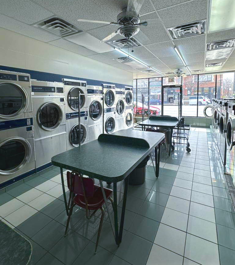 Empty laundromat with washing machines along the walls, tables and chairs in the center, and large windows showing parked cars outside.