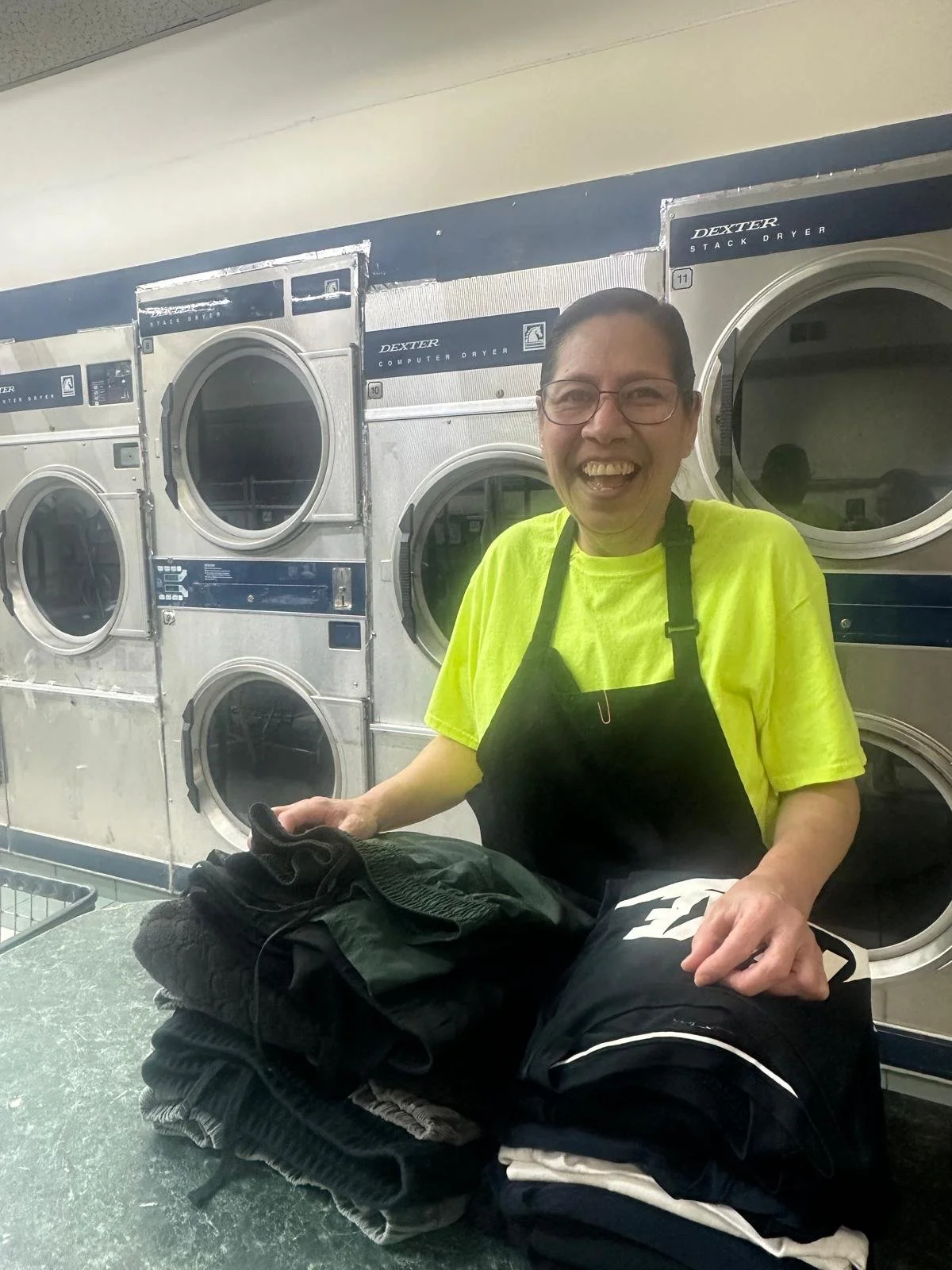 A woman with glasses wearing a bright yellow shirt and black apron, smiling at the camera, is folding laundry at a laundromat. There are stacked washing machines behind her.