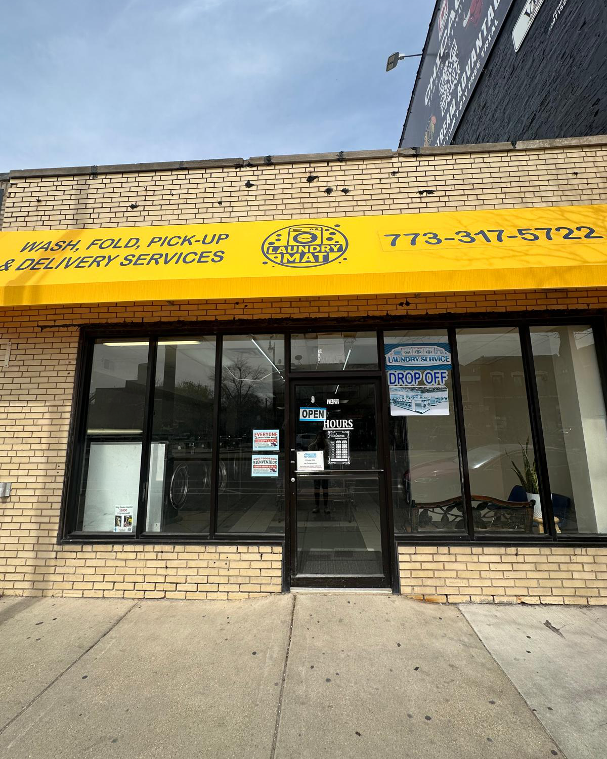 Front view of a laundromat with a yellow sign that reads "Wash, Fold, Pick-Up & Delivery Services" and includes the business name "Laundry Mat" and phone number 773-317-5722. The entrance has glass doors and large windows, with signs indicating "Open," "Hours," and "Drop Off." Inside, laundry machines are visible.