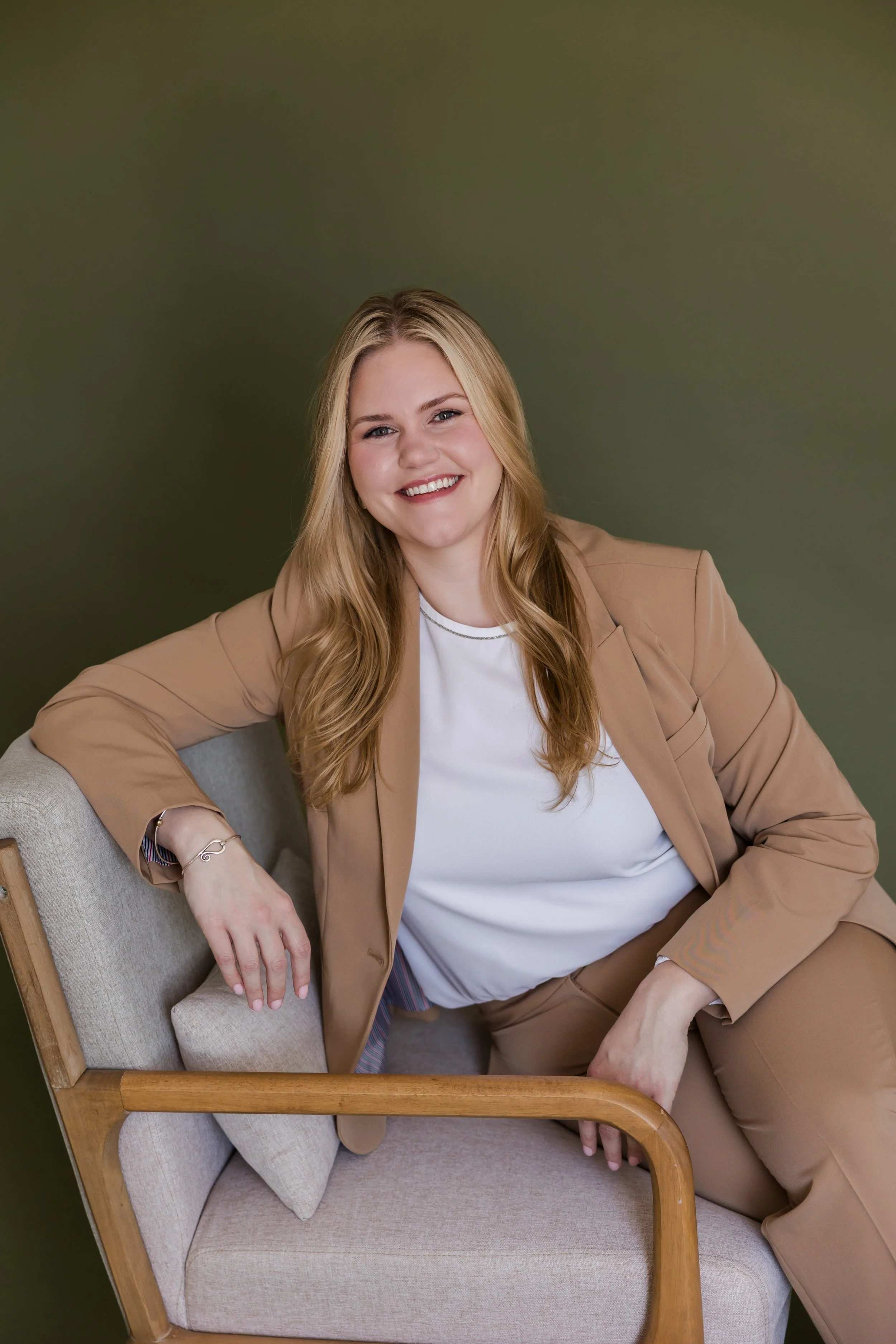 A woman with long blonde hair, smiling, sitting on a light-colored armchair in front of a plain green wall, wearing a beige blazer and pants with a white t-shirt.