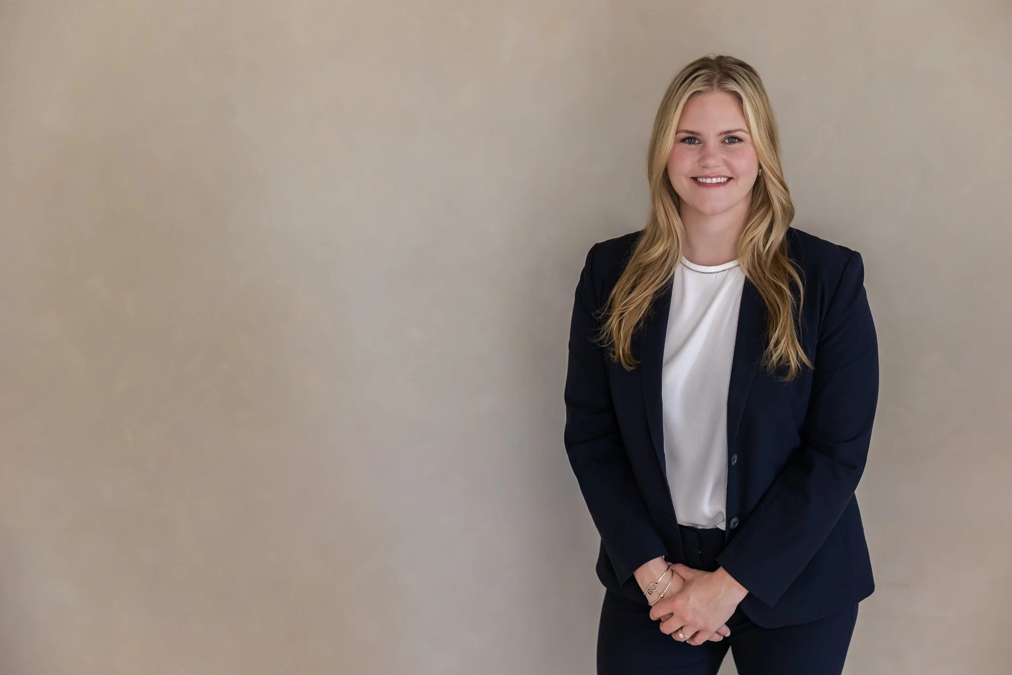 A young woman with long blonde hair smiling, dressed in a white top and navy blazer, standing against a plain beige wall.