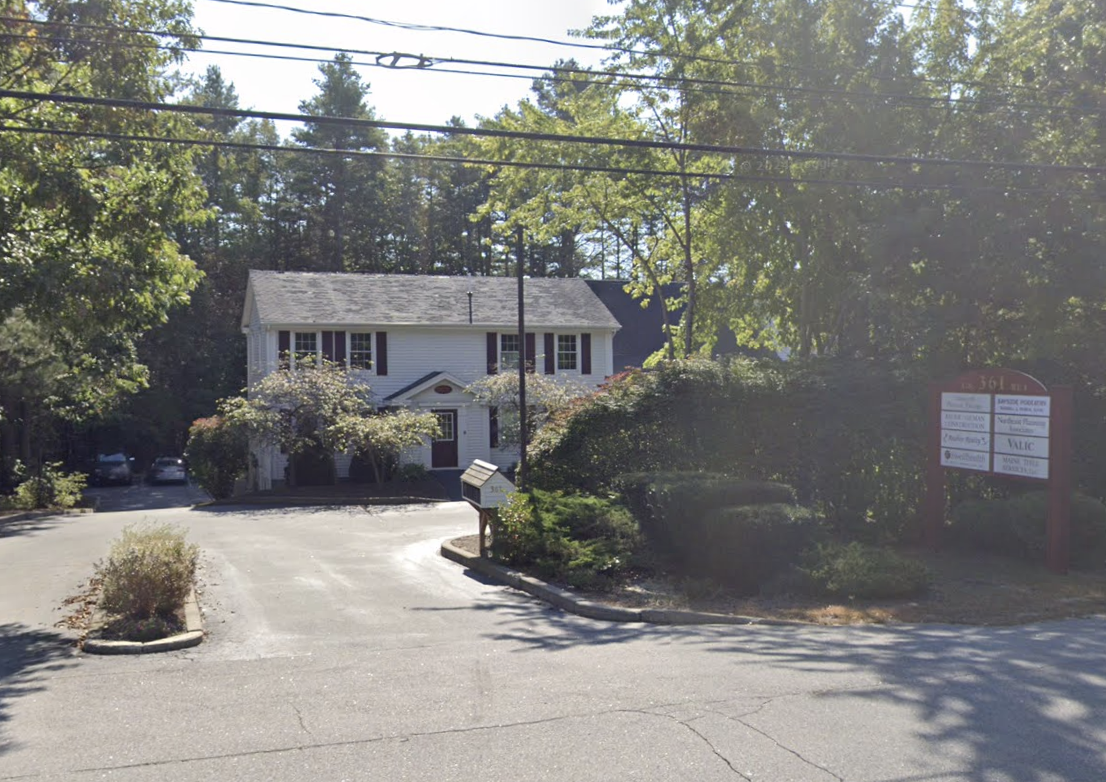 A white two-story house with dark shutters, surrounded by trees and shrubs, with a gravel driveway and a small mailbox in the front yard. There is a sign on the right side.