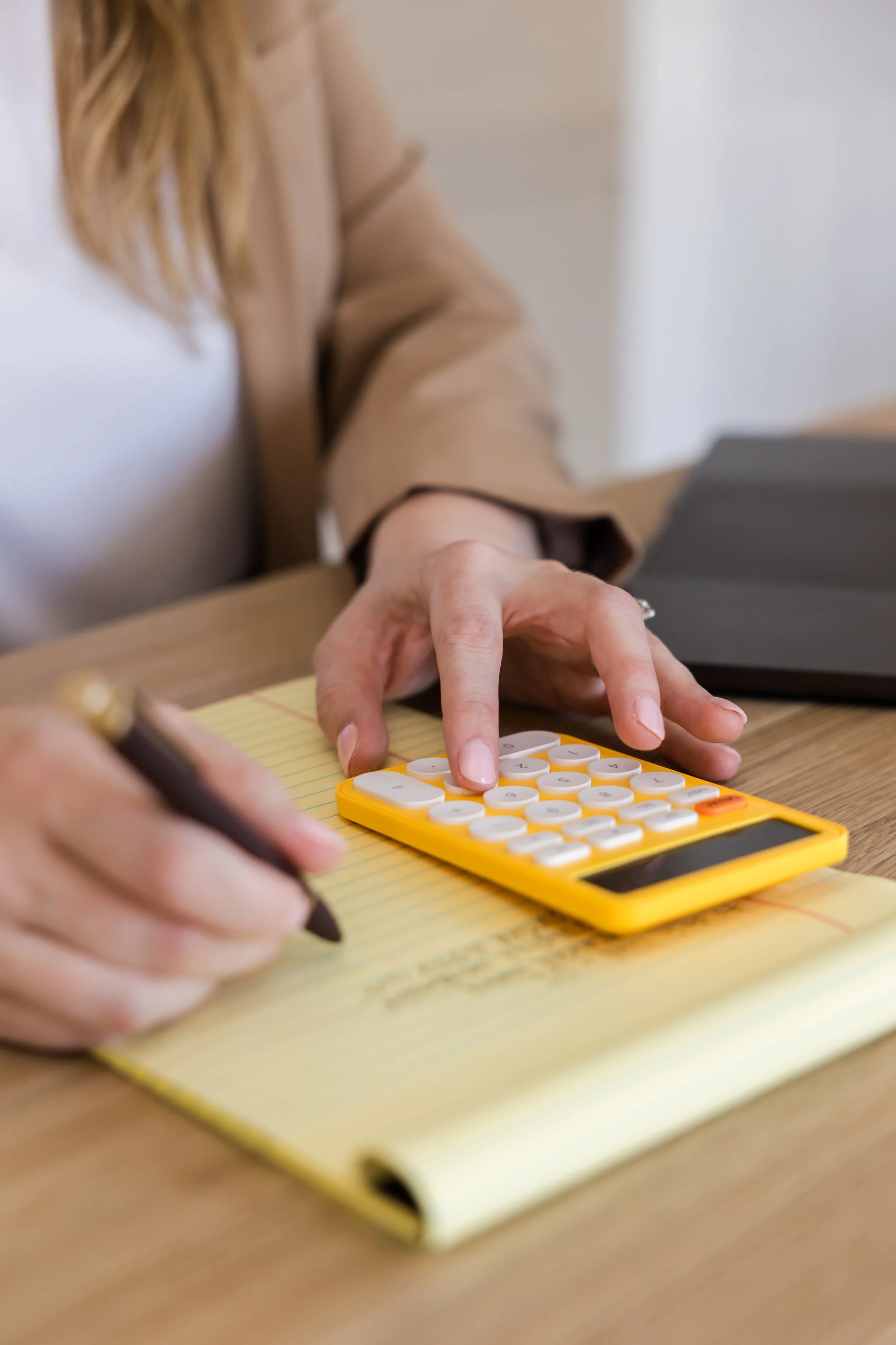 A person is pressing buttons on a yellow calculator while sitting at a wooden desk, with a pen in their left hand and a yellow notepad in front of them.