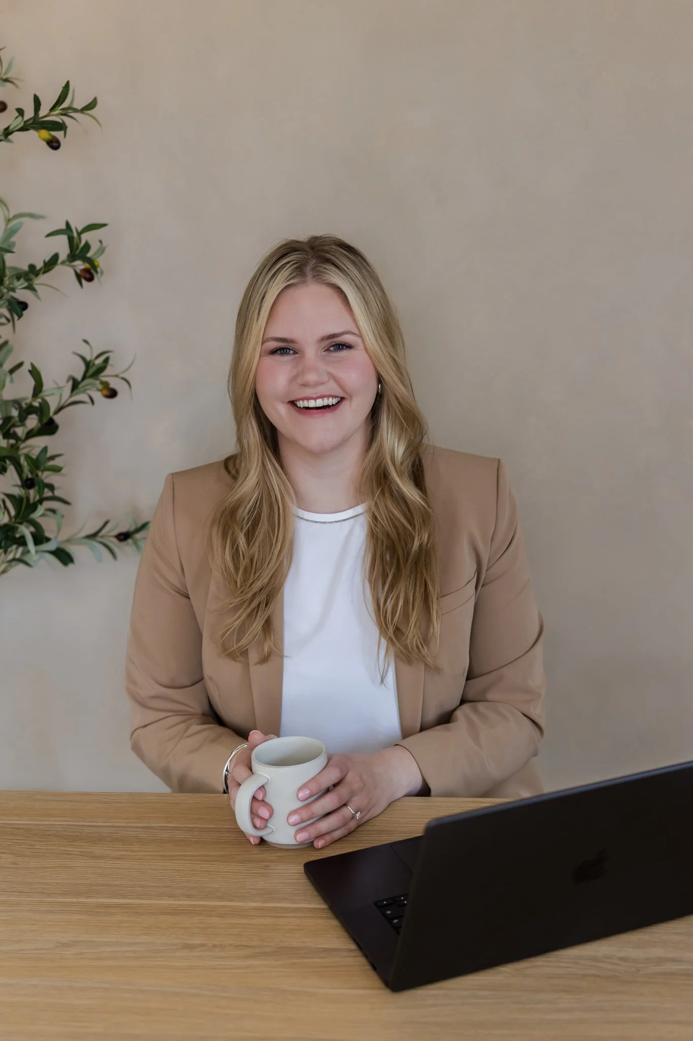 A smiling woman with blonde hair and a tan blazer sitting at a wooden desk, holding a white mug, with a laptop in front of her and a potted plant in the background.