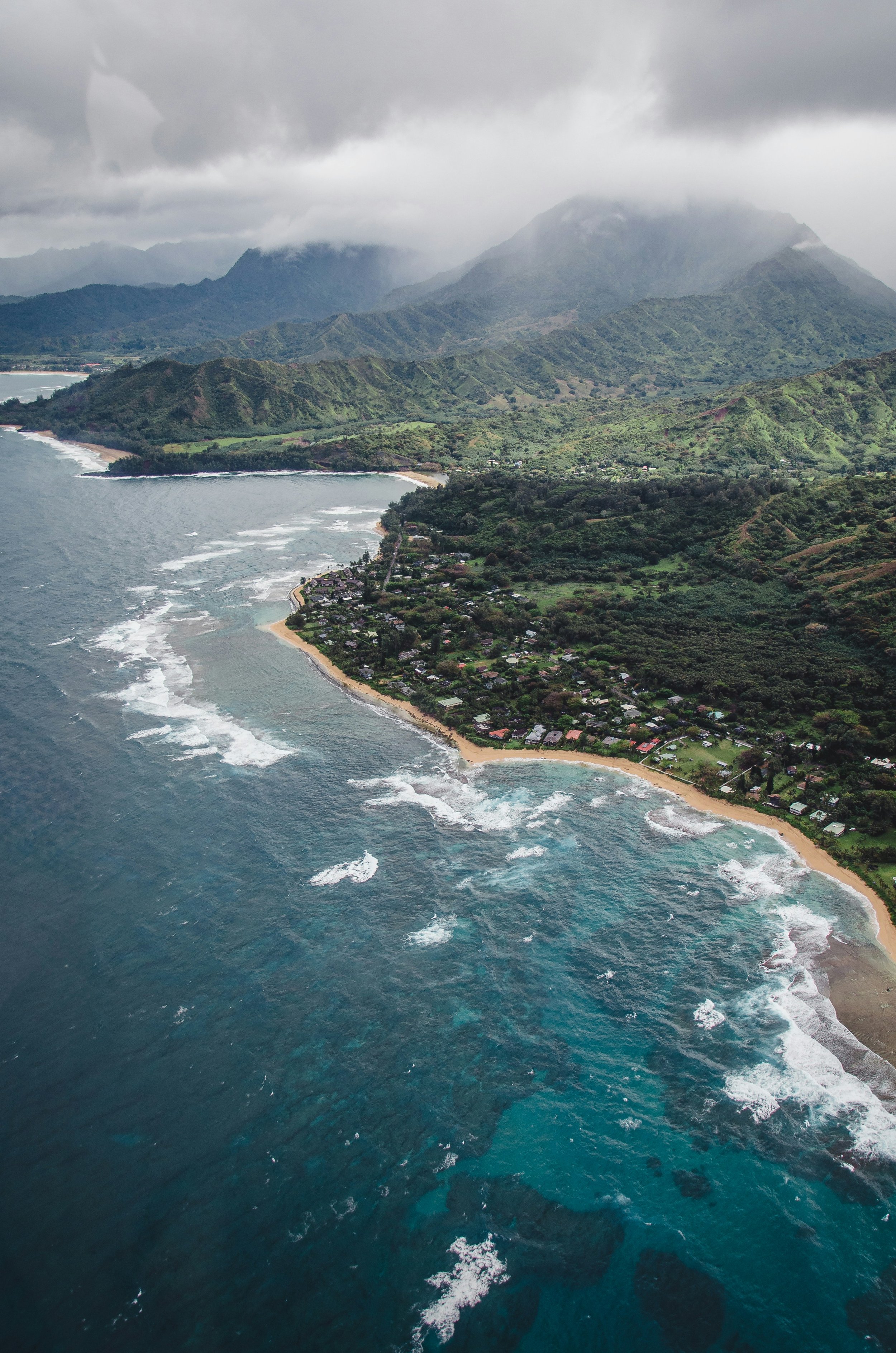 Aerial view of a coastal area with a beach, residential houses, lush green hills, and mountains in the background under cloudy skies.