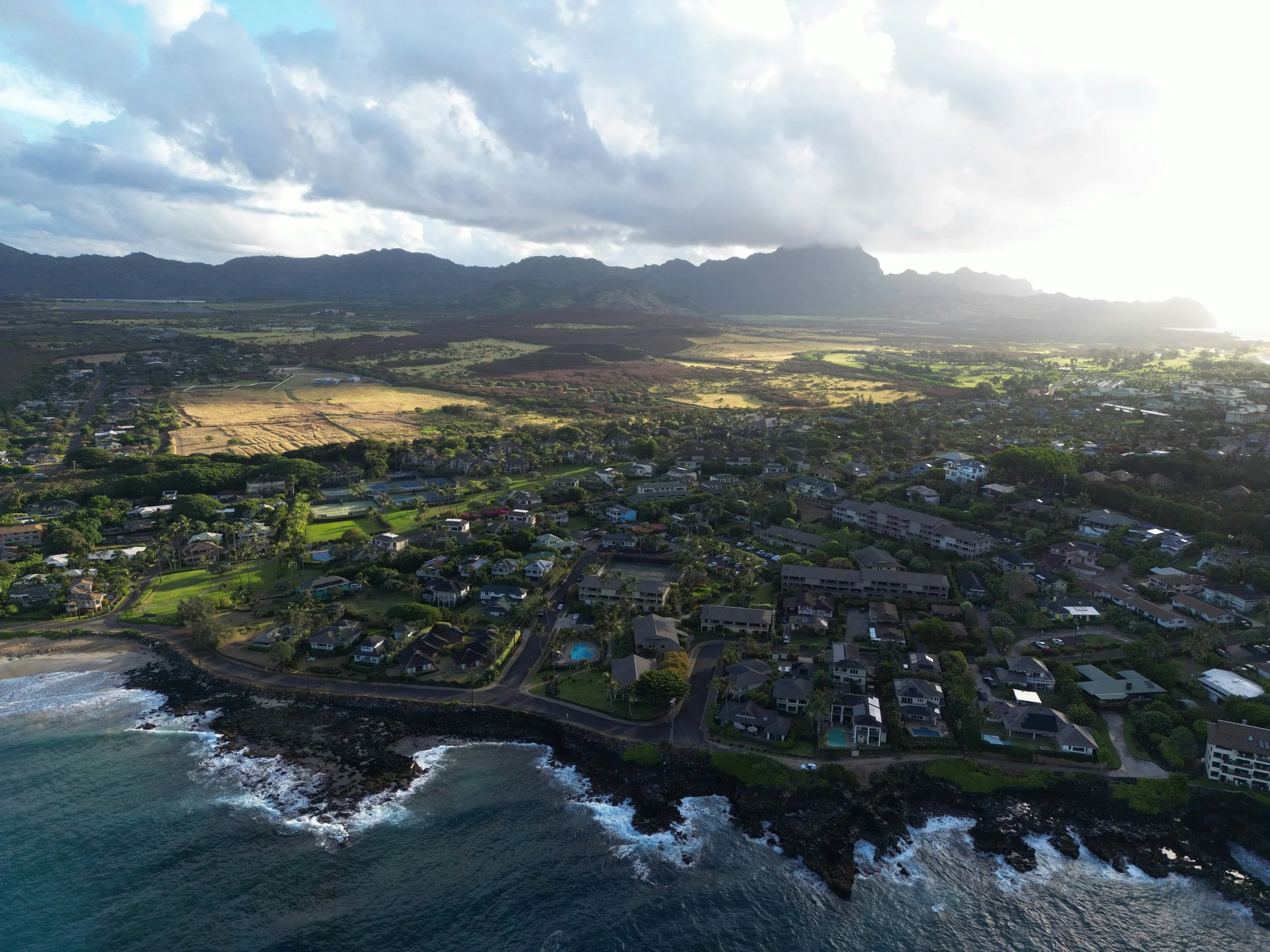 Aerial view of a coastal residential area with houses, roads, a coastline, and ocean waves, with hills and mountains in the background, and partly cloudy sky.