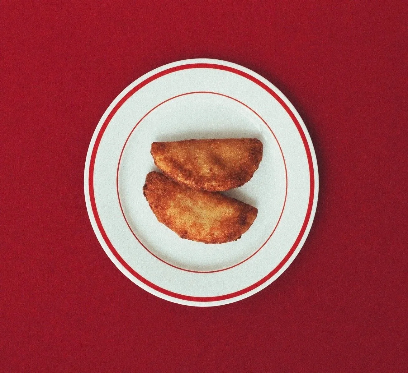 Handmade Portuguese rissois by Rissolá, featuring two golden, crispy patties on a white plate against a red background.