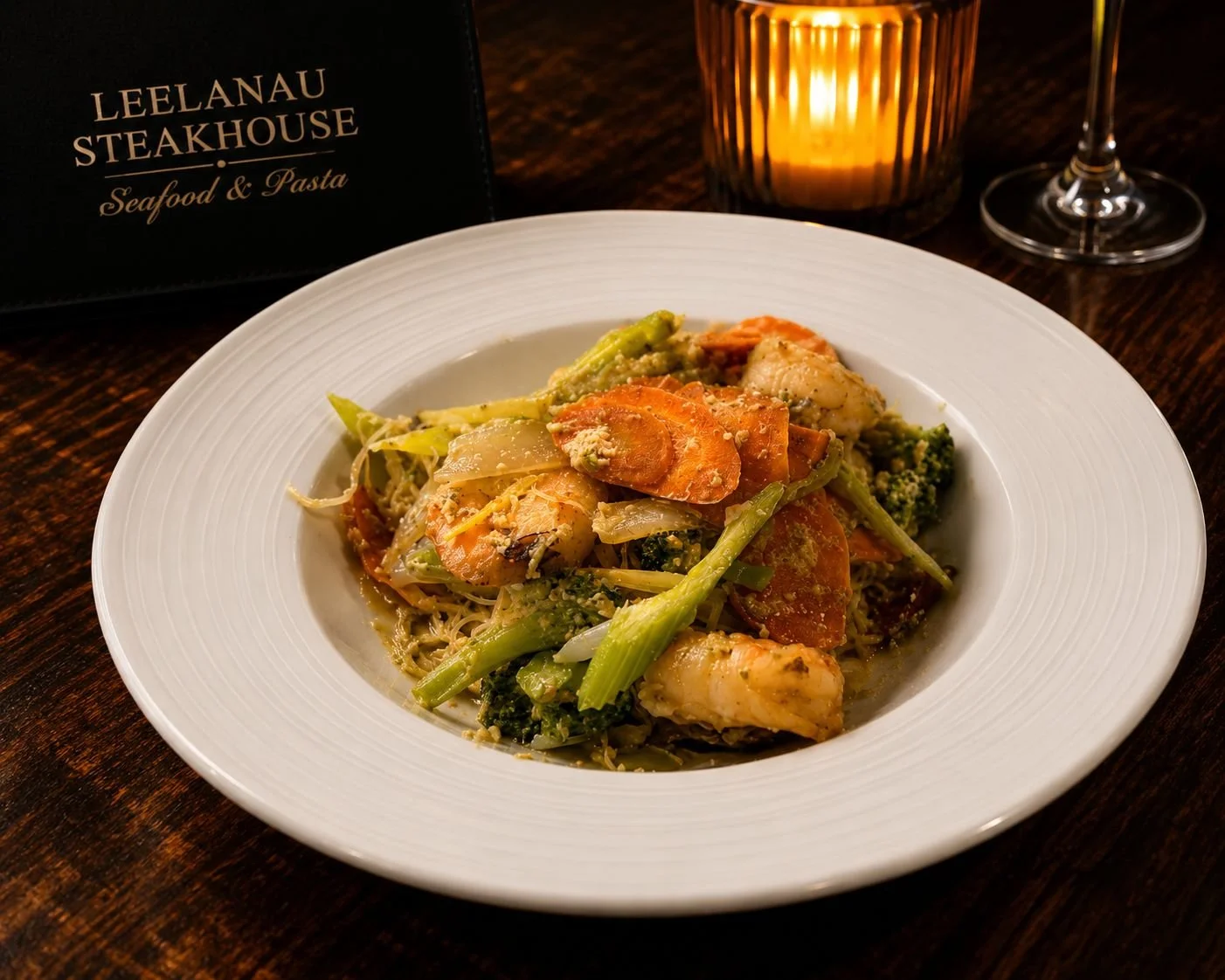A plate of seafood and pasta with vegetables, served at Leelanau Steakhouse, with a candle and a glass of wine in the background.