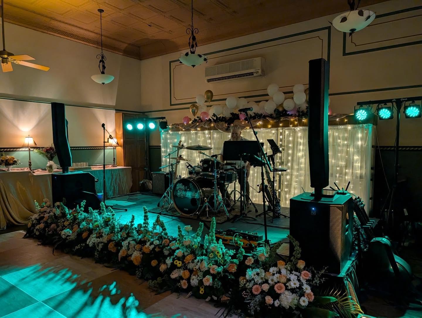 Indoor event with a decorated stage, musical instruments, floral arrangements, and ambient lighting, likely for a celebration or wedding reception.