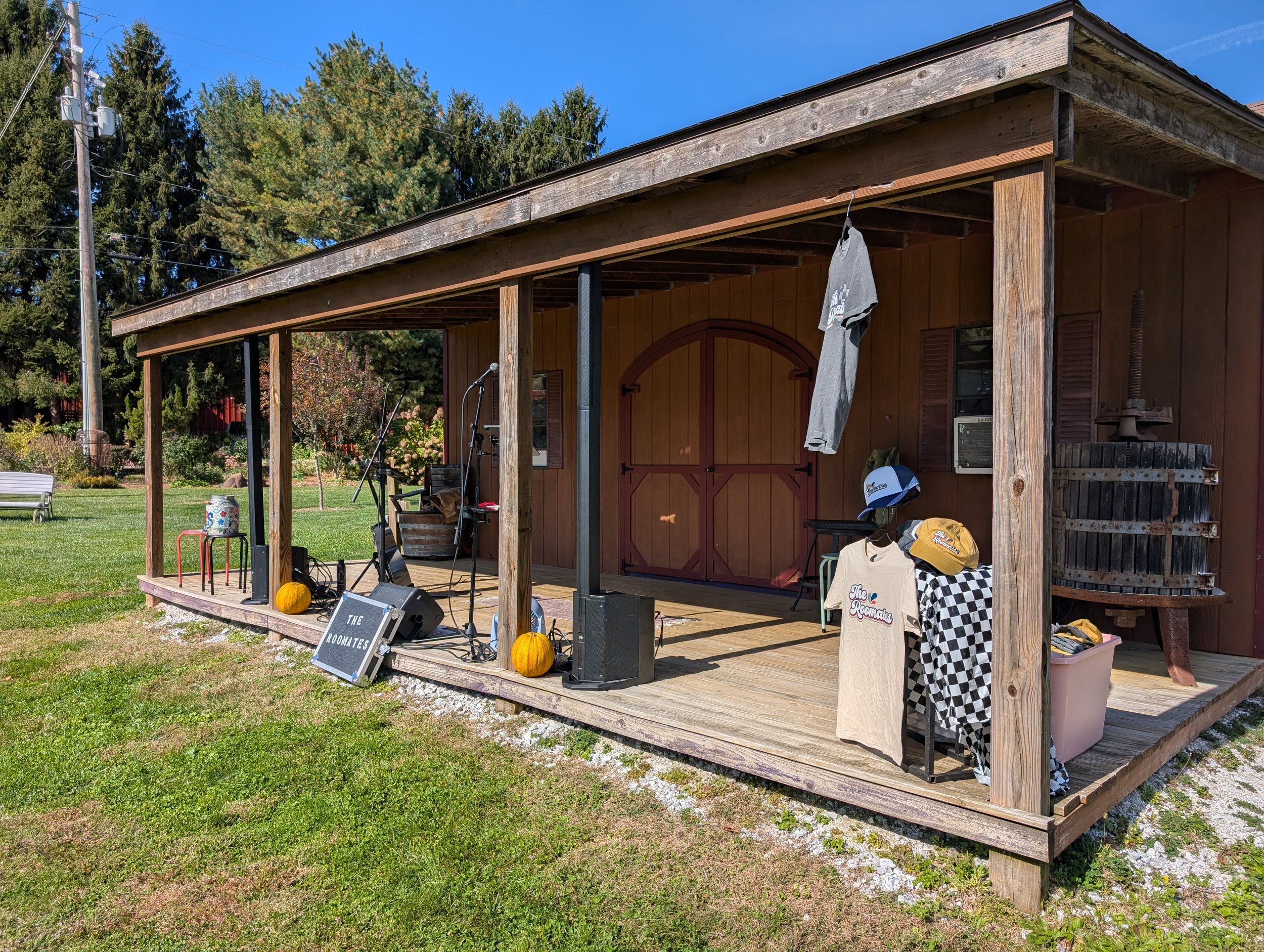 A wooden porch with string lights, a microphone, and various items including pumpkins, clothes, and a sign reading 'The Roomates,' with a garden, trees, and a blue sky in the background.