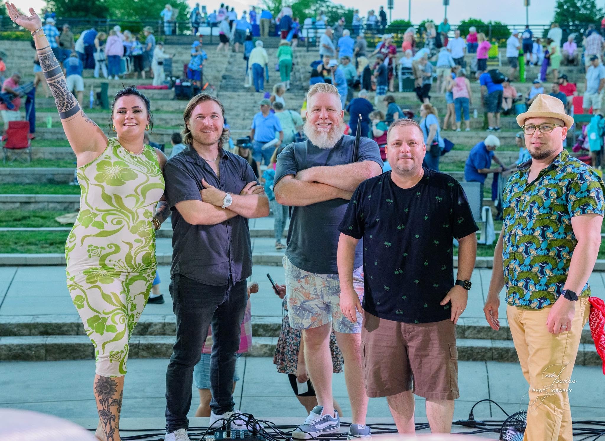 Five people standing outdoors at an event with many people on steps behind them. The group includes a woman with dark hair in a green floral dress with tattoos on her arm, a man with medium length hair in a dark shirt, a bearded man with short light hair in a dark shirt, a man with short hair in a black shirt, and a man with glasses, a tan hat, and a colorful Hawaiian shirt.
