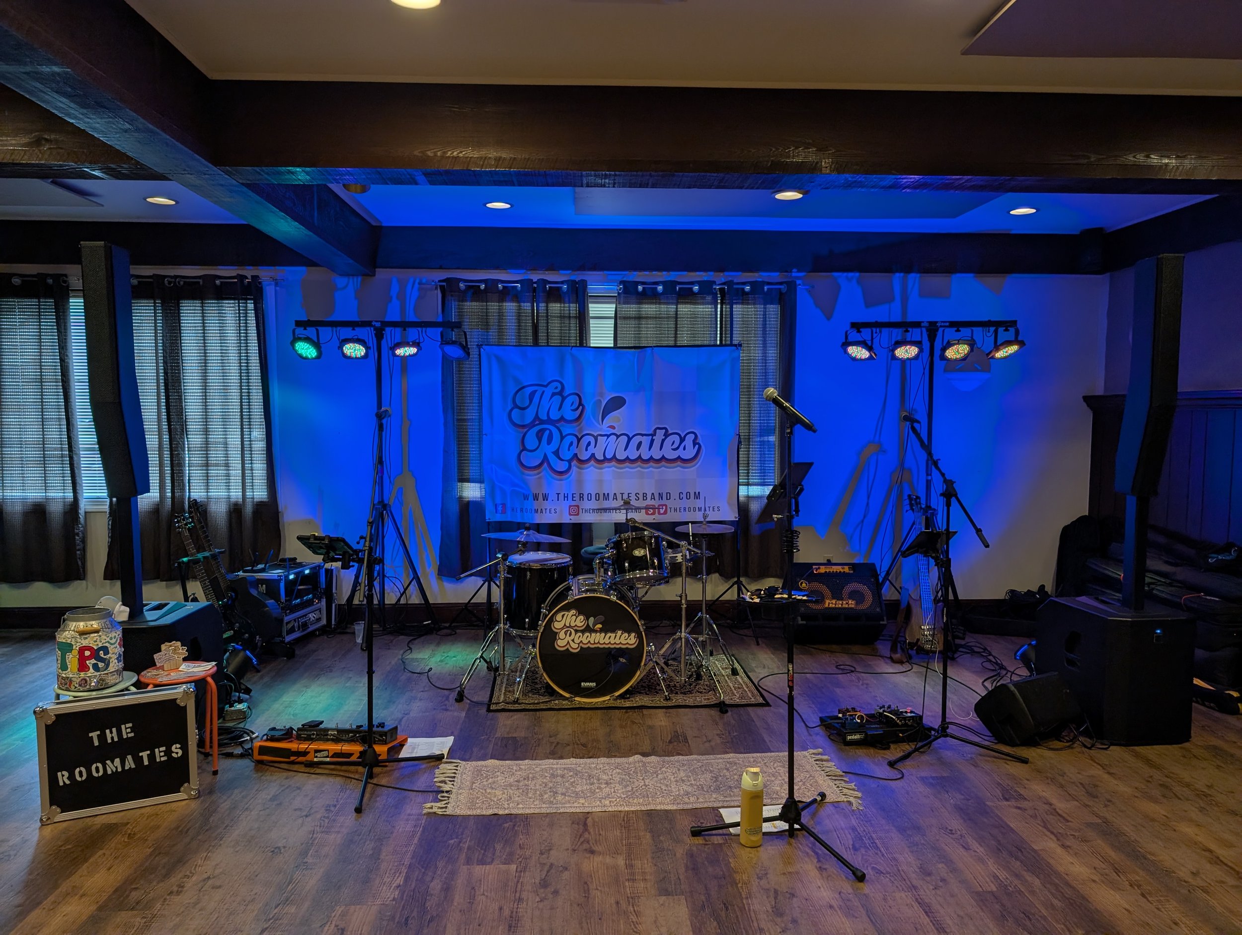 Stage setup with drum kit, microphones, lighting, and a banner for The Roomates band, in a cozy indoor venue with curtains and wooden floors.