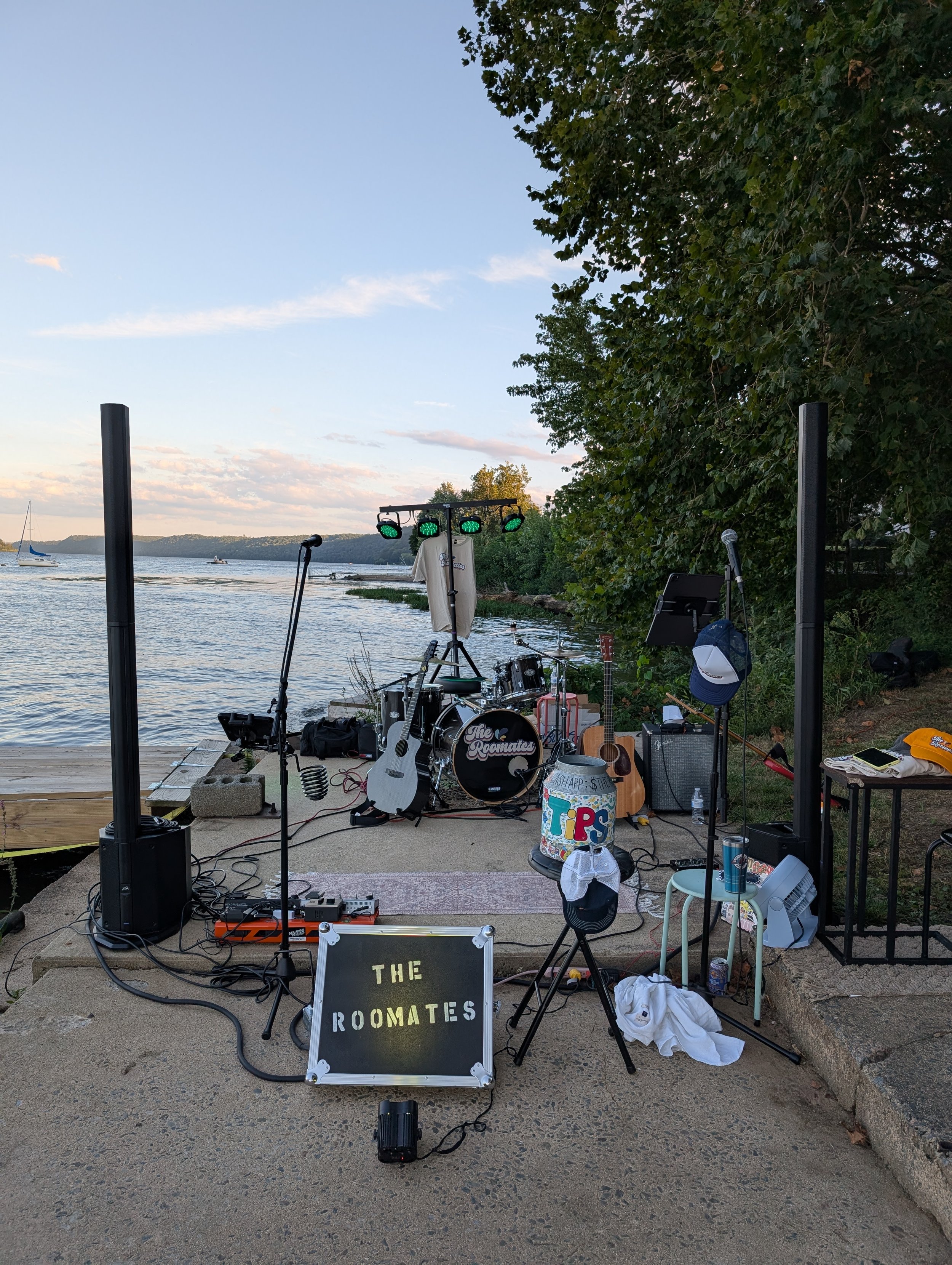 Outdoor scene by a water with a small band setup including drums, guitars, microphone, speakers, and music equipment, set against a backdrop of trees and water, with a sign that says 'The Roomates'.