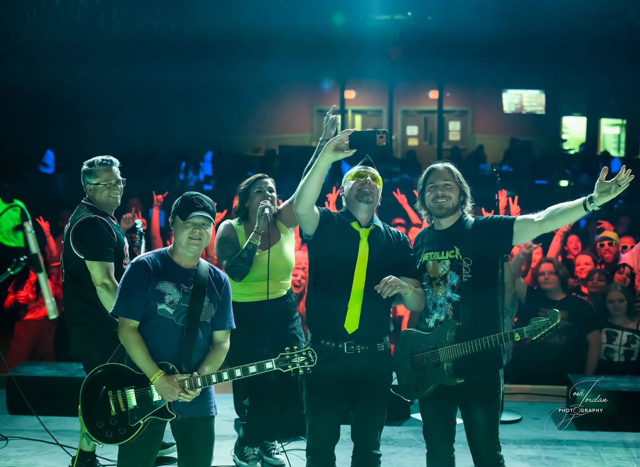 A group of people standing indoors, some with guitars, taking a selfie or photo together. The background includes a crowd, some in bright clothing, and a stage setting with lighting.