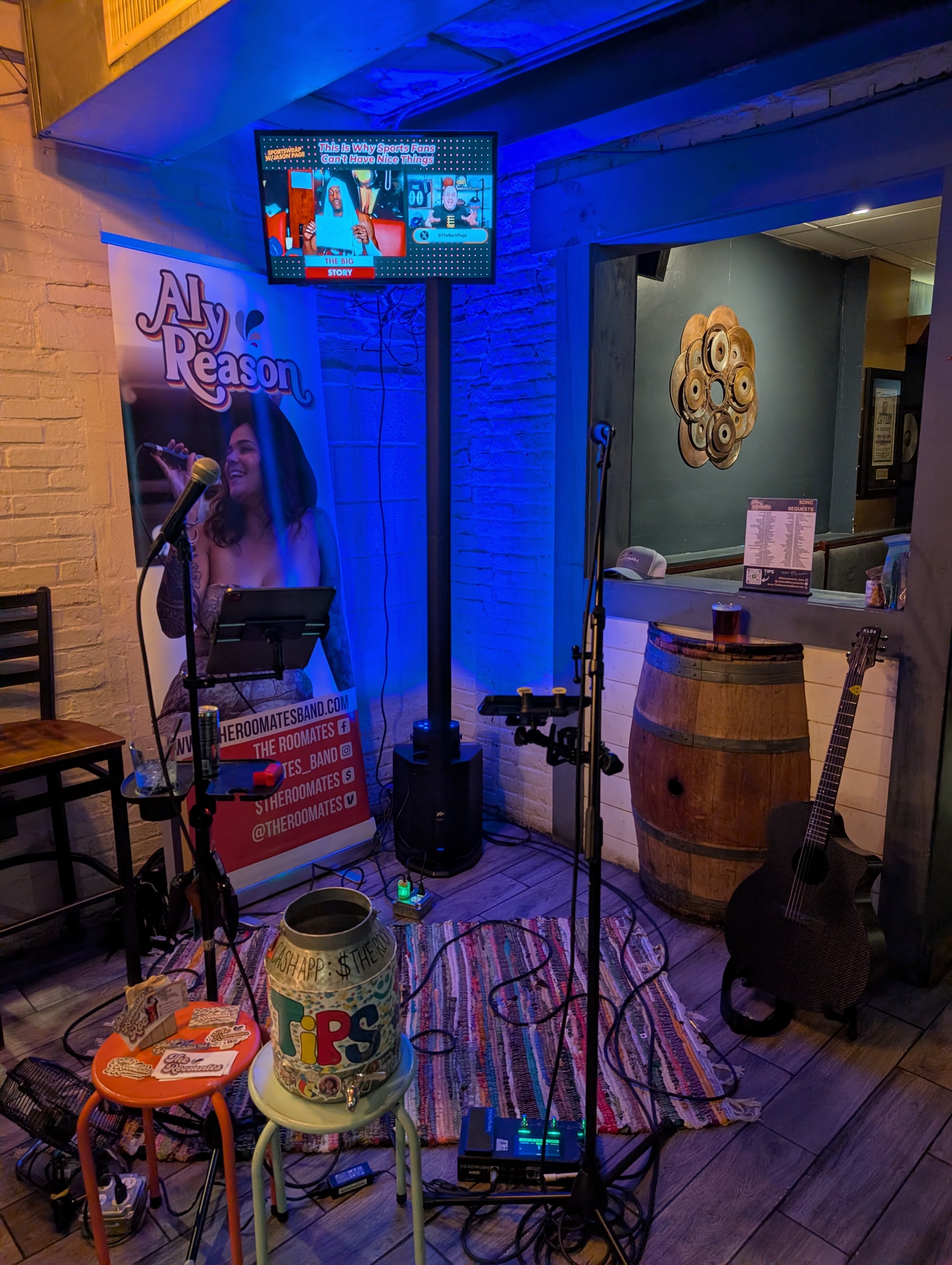 A small stage setup in a bar with two microphones, a guitar, and various lighting and tech equipment. There is a colorful poster on the left with a woman singing and promotional text. A TV screen displays a news segment about sports fans. The bar has a rustic decor with a barrel used as a table, a decorative wall art piece, and a menu on the counter.