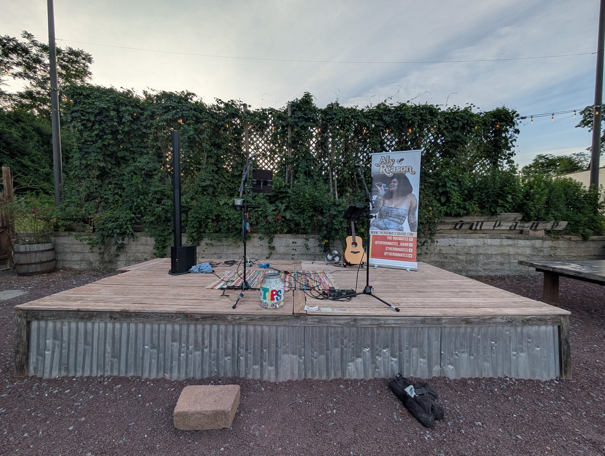 An outdoor stage set up with a microphone, guitar, and sound equipment, with a banner of a woman singing, and surrounded by greenery and string lights.