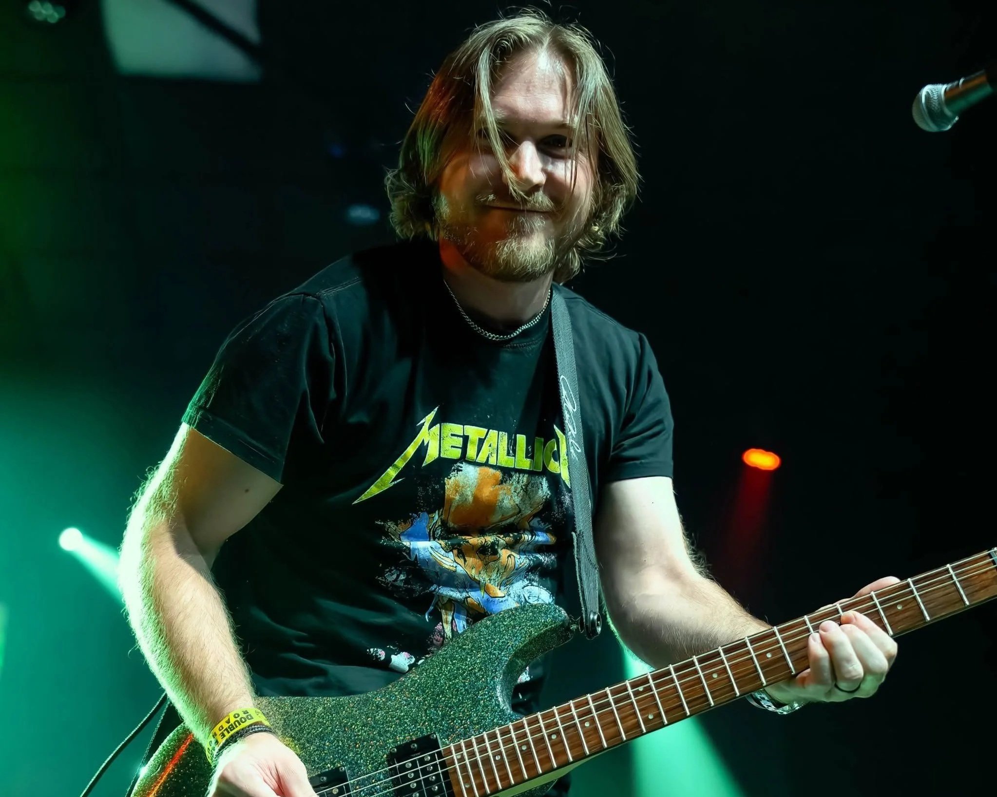 A man with long hair and a beard playing an electric guitar on stage, wearing a Metallica t-shirt and smiling.