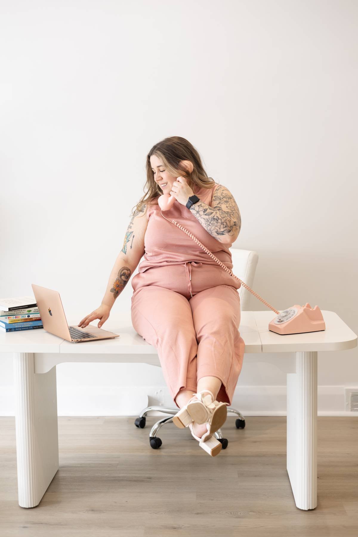 A woman in pink lounge wear sitting on a white desk, talking on a pink rotary phone, with a laptop and books nearby, smiling and looking down.