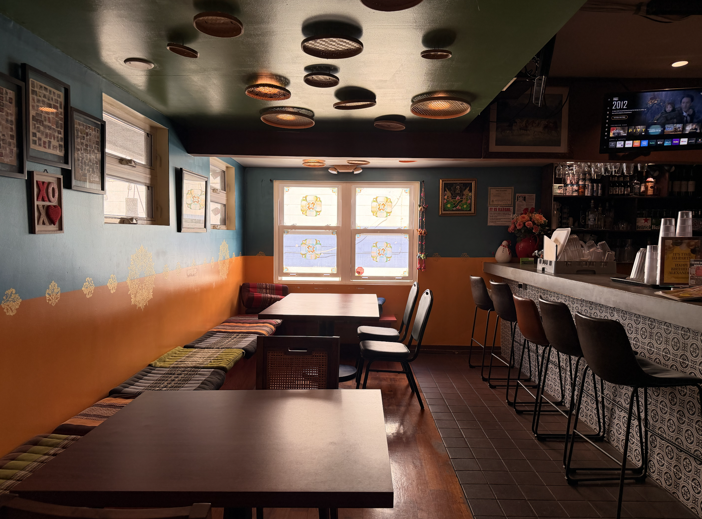 The image shows the interior of our cozy restaurant with different colored walls, Thai-patterned cushions, and wooden tables.