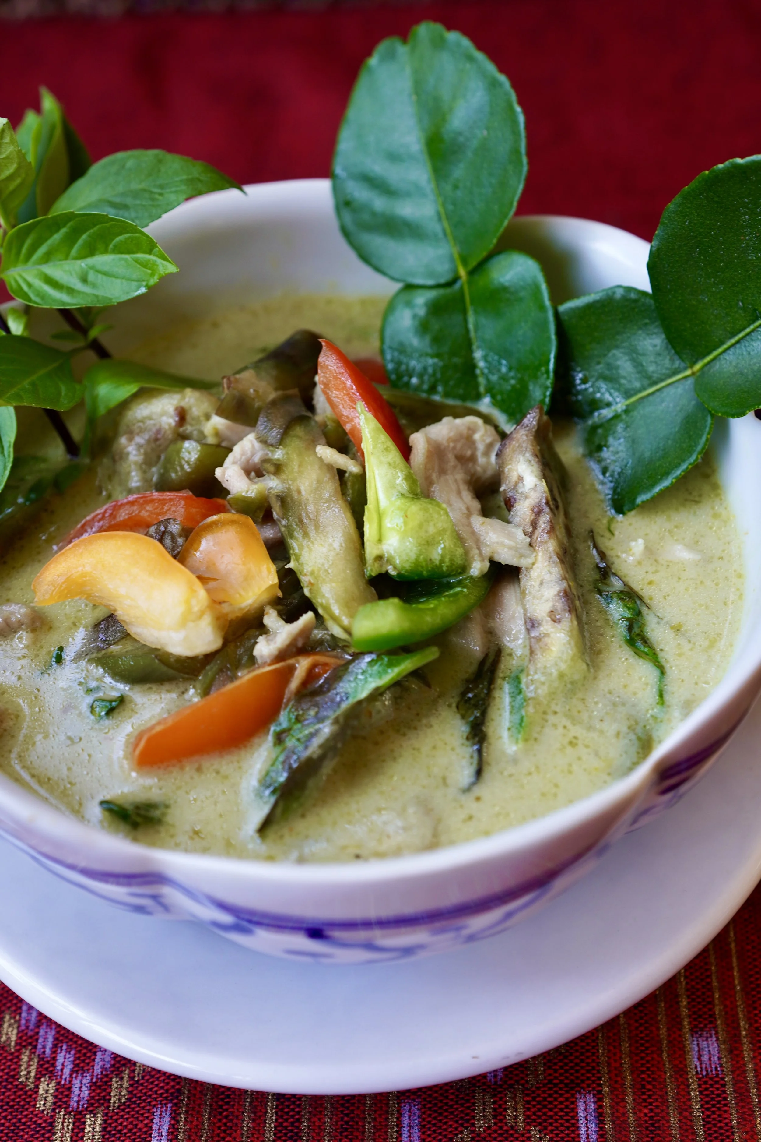 A bowl of green curry with vegetables and herbs on a red woven placemat.