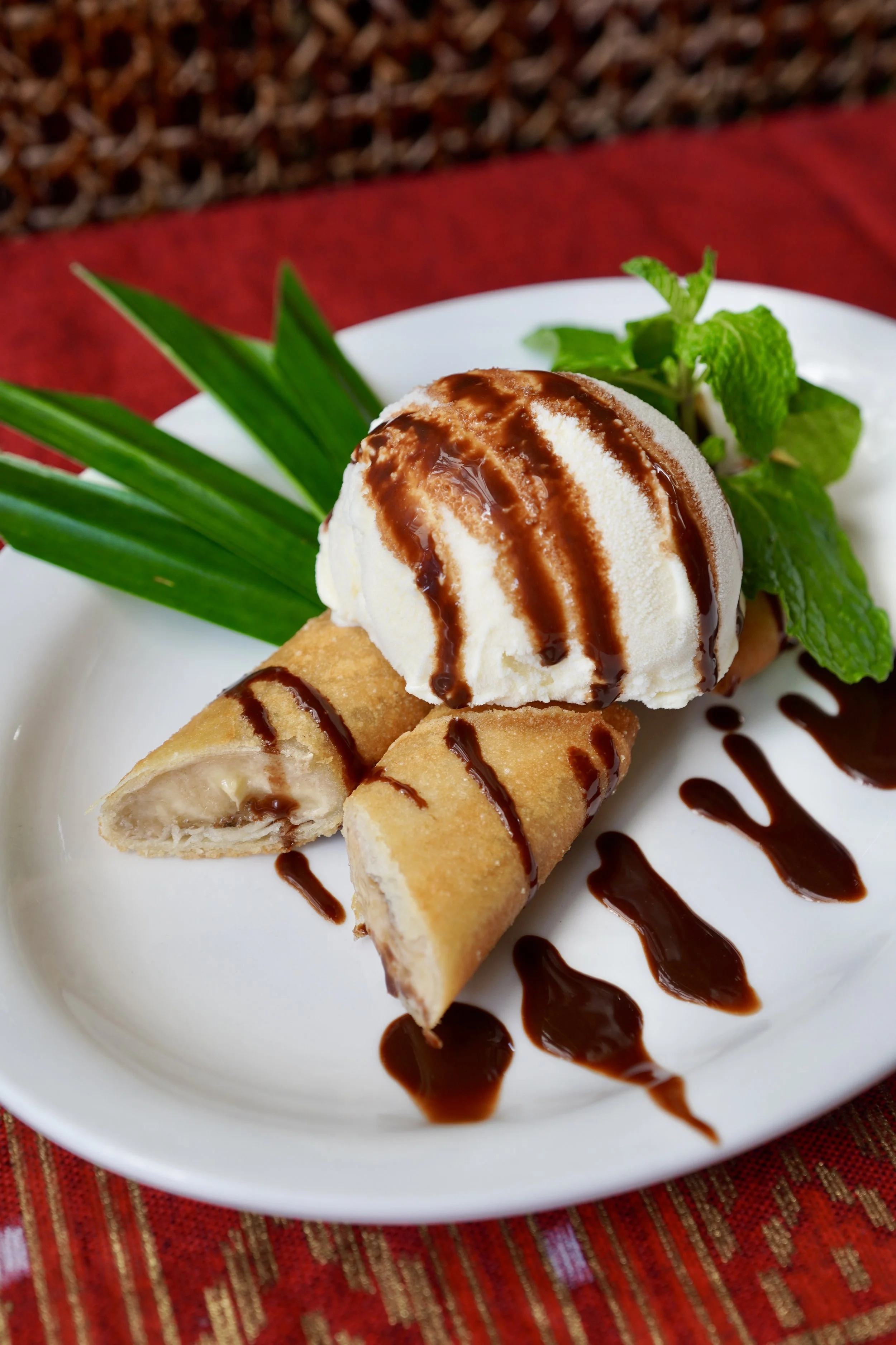 Banana spring rolls with vanilla ice cream, chocolate drizzle, and mint garnish on a white plate, set on a red tablecloth.