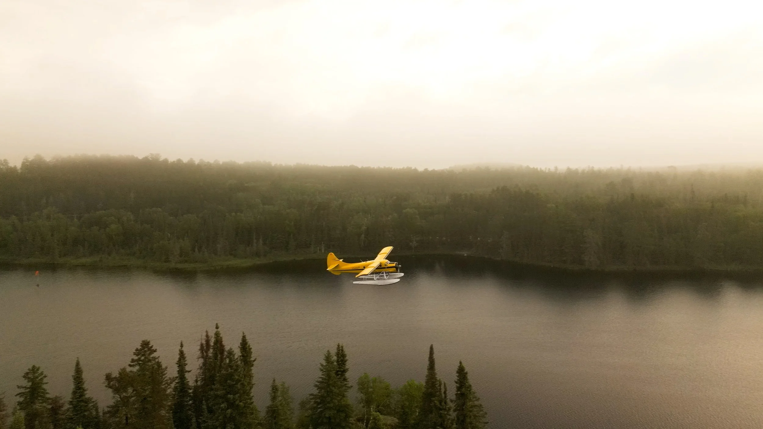 Yellow floatplane flying over a body of water with a forested shoreline under a cloudy sky.