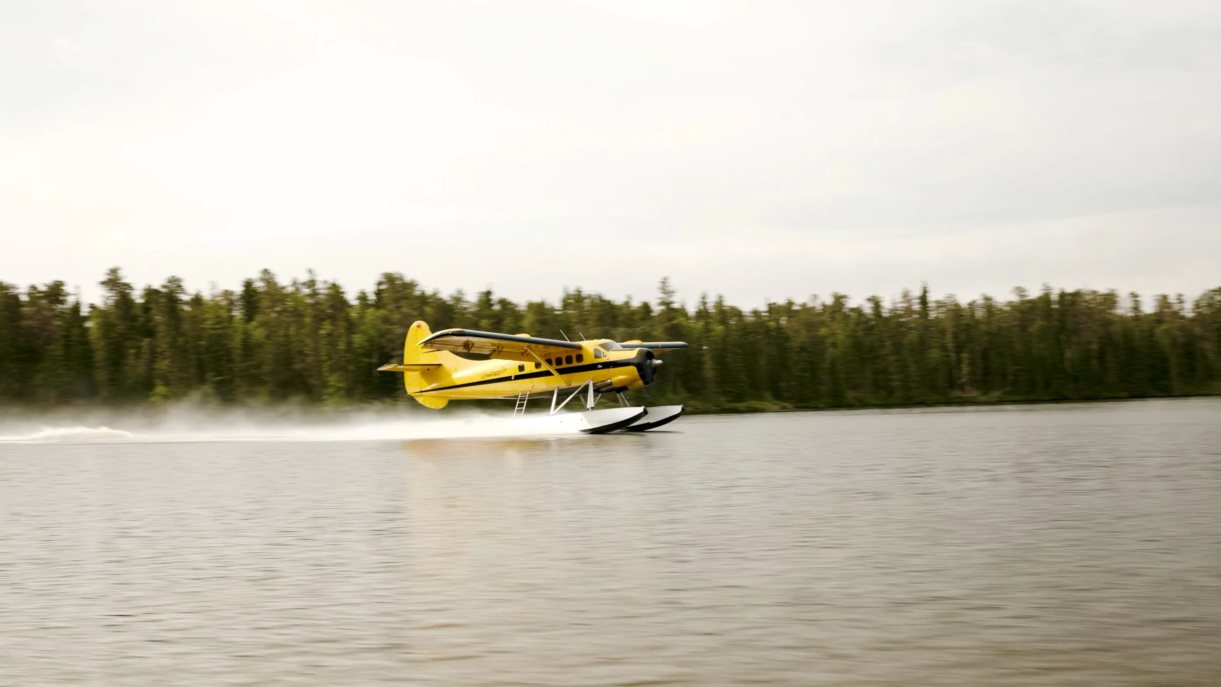 A yellow seaplane taking off from a body of water, creating a spray of water behind it, with a forested shoreline in the background.