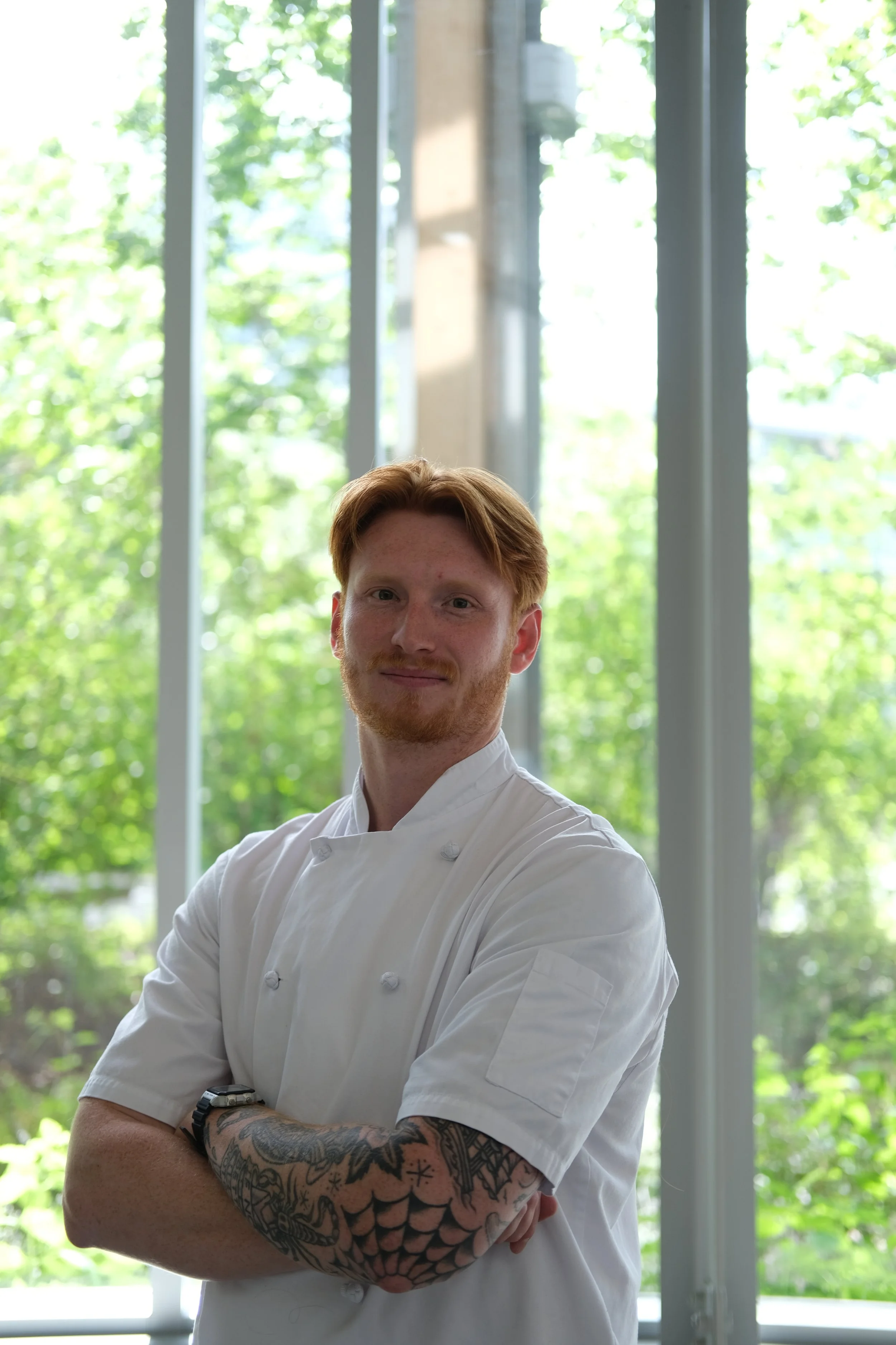 A male chef with red hair and a beard, wearing a white chef's coat, standing with arms crossed in front of large windows with green trees outside.
