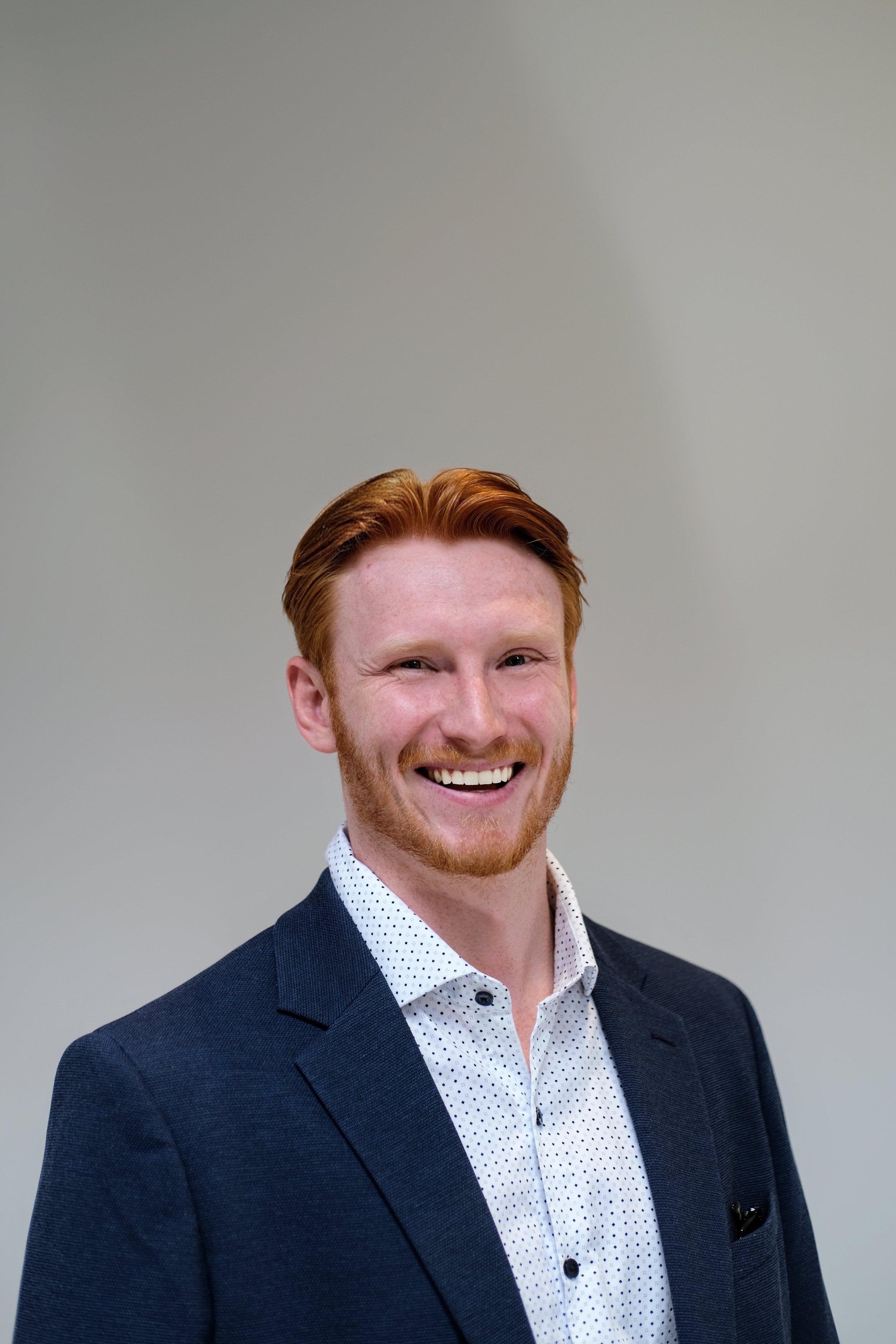 Portrait of a smiling man with red hair and beard wearing a dark suit and white polka dot shirt against a plain gray background.