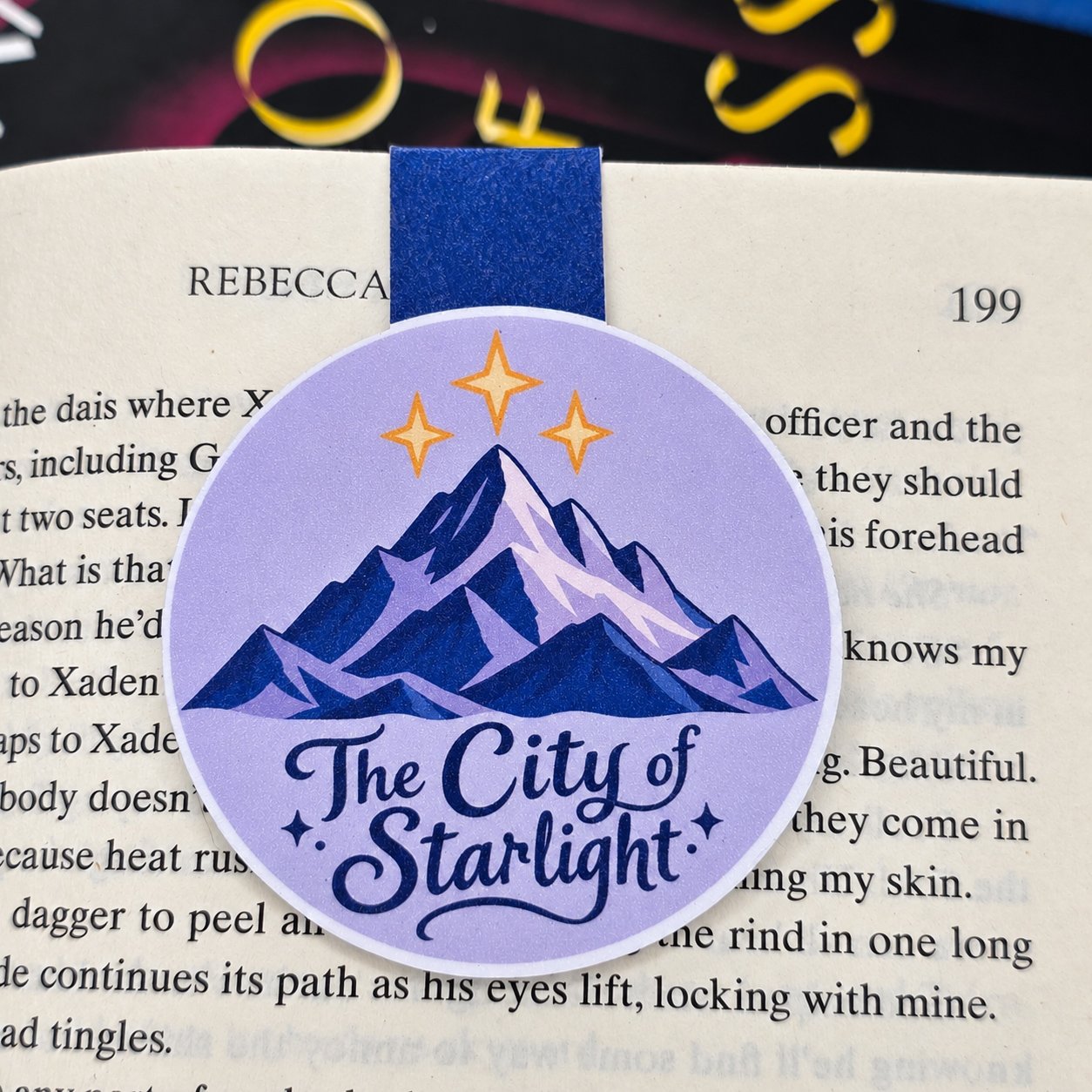 Round double-sided magnetic bookmark featuring blue mountains, three gold stars, and “The City of Starlight” lettering on lavender.