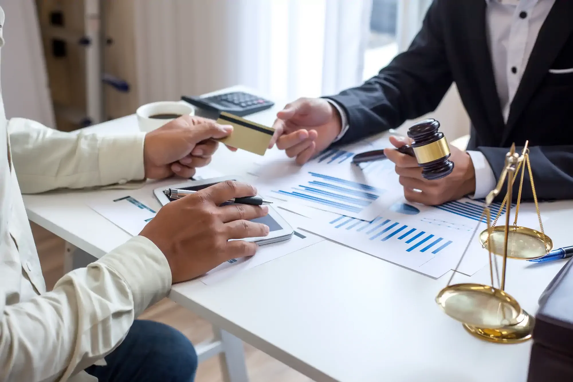 Una reunión de negocios entre dos personas en la que se intercambia un libro o documento. Sobre la mesa hay gráficos financieros, un mazo, una calculadora, una tableta, una taza de café y una balanza dorada.