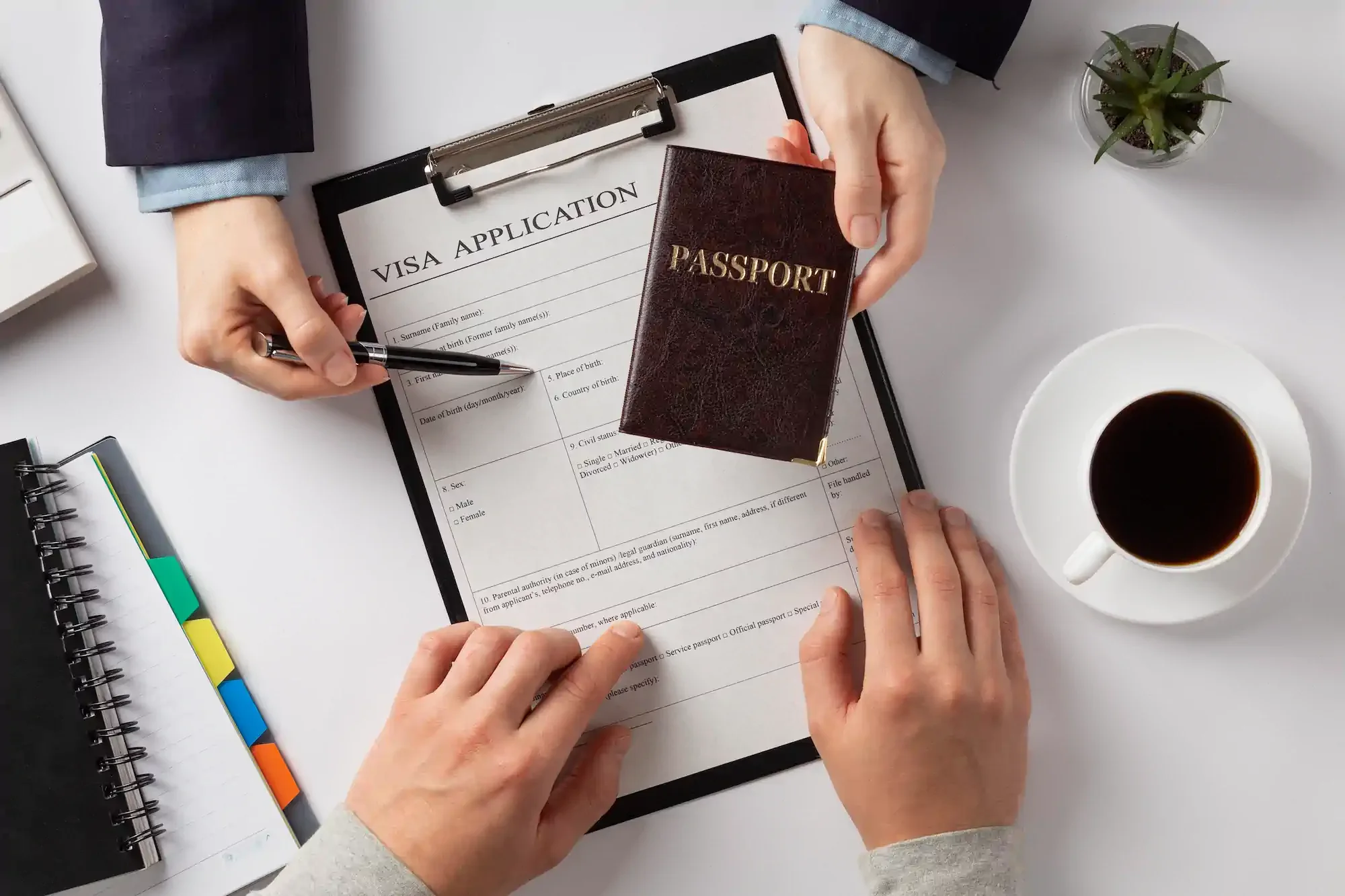 Una persona rellena un formulario de solicitud de visado sobre una tablapapeles con un bolígrafo; sostiene un pasaporte en la mano izquierda, y hay una taza de café sobre una mesa blanca. En la mesa también se encuentran una pequeña planta en maceta,
