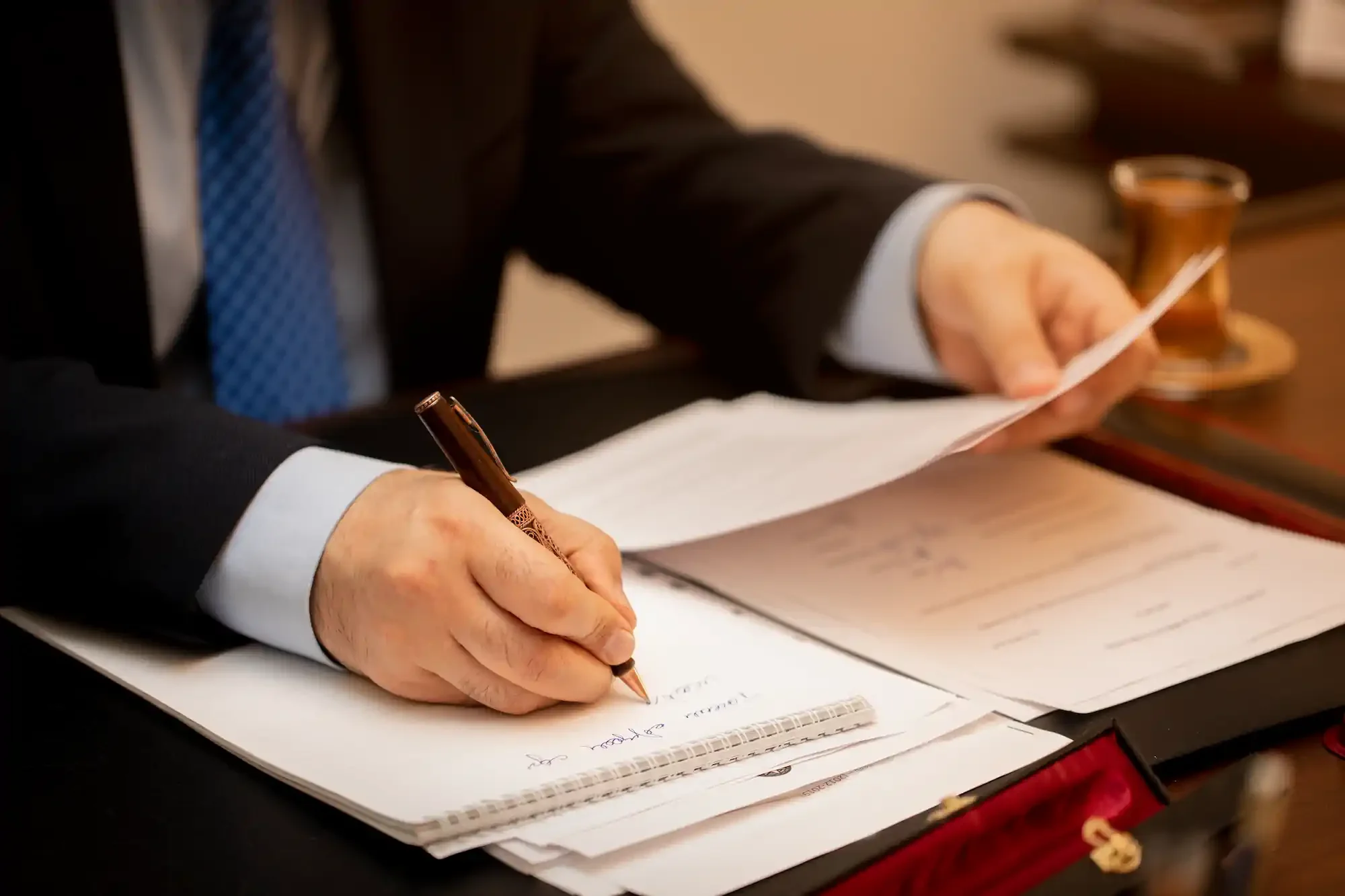 Una persona con traje negro y corbata azul está firmando documentos con un bolígrafo dorado, mientras apoya la mano izquierda sobre el papel.