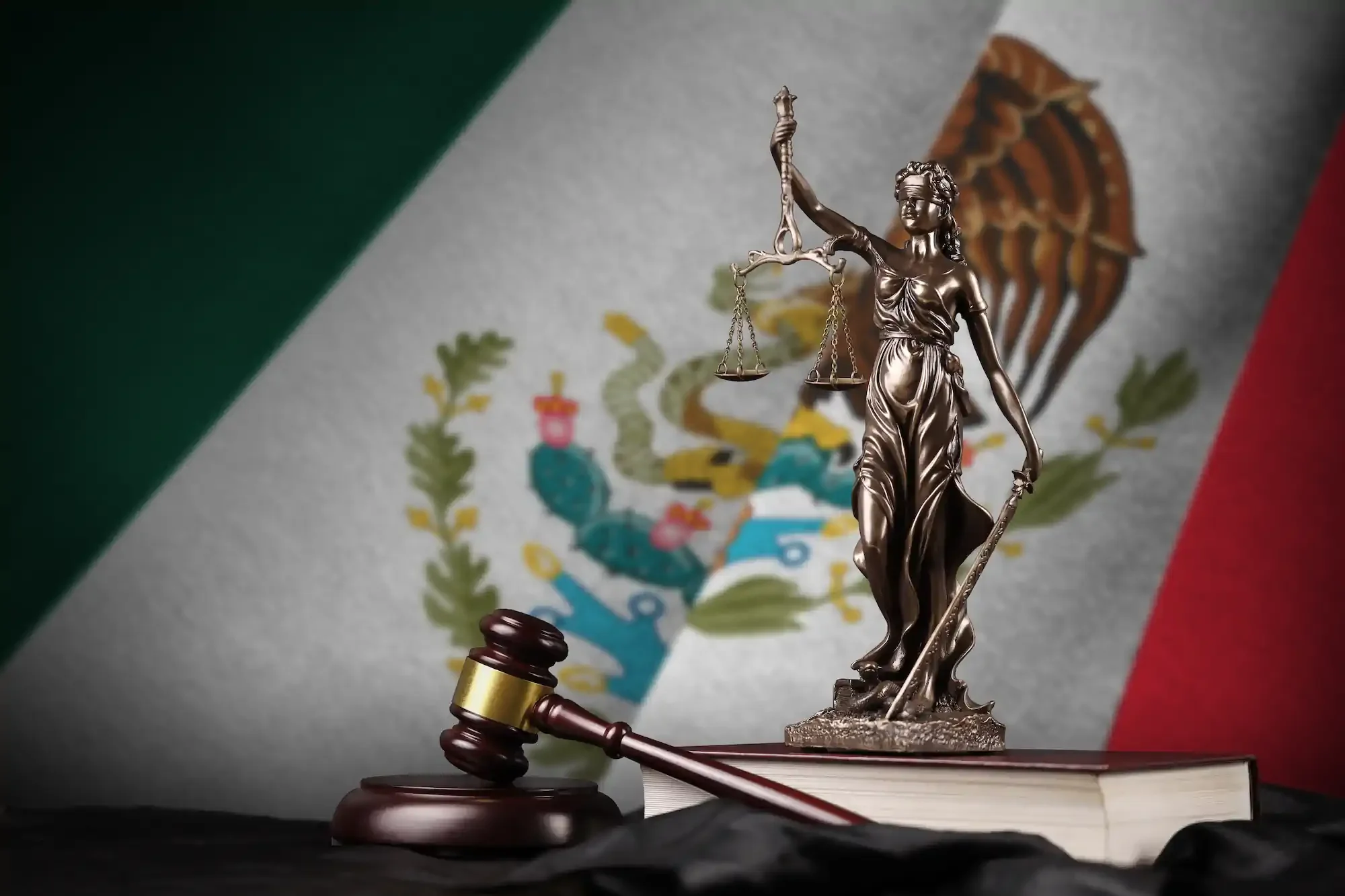 Estatua de la Justicia que sostiene una balanza y una espada, con un mazo y un libro jurídico delante, y una bandera de México con su escudo al fondo.