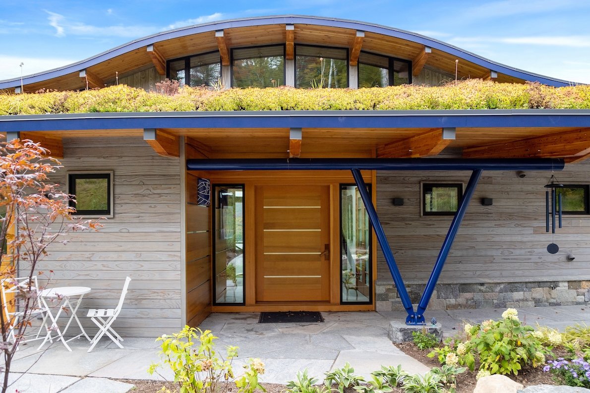 Entry to a custom home in Wilmington, Vermont, with a planted roof, warm wood siding, and a broad overhang supported by angled steel braces.