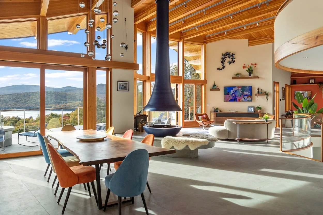 Open dining and living area in a custom home in Wilmington, Vermont, with a suspended fireplace, wood ceilings, and expansive windows overlooking the lake and mountains.