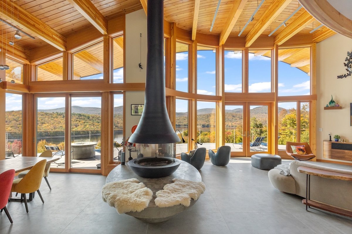 Living area in a custom home in Wilmington, Vermont, with a suspended fireplace, soaring wood ceilings, and floor-to-ceiling windows overlooking the lake and mountains.