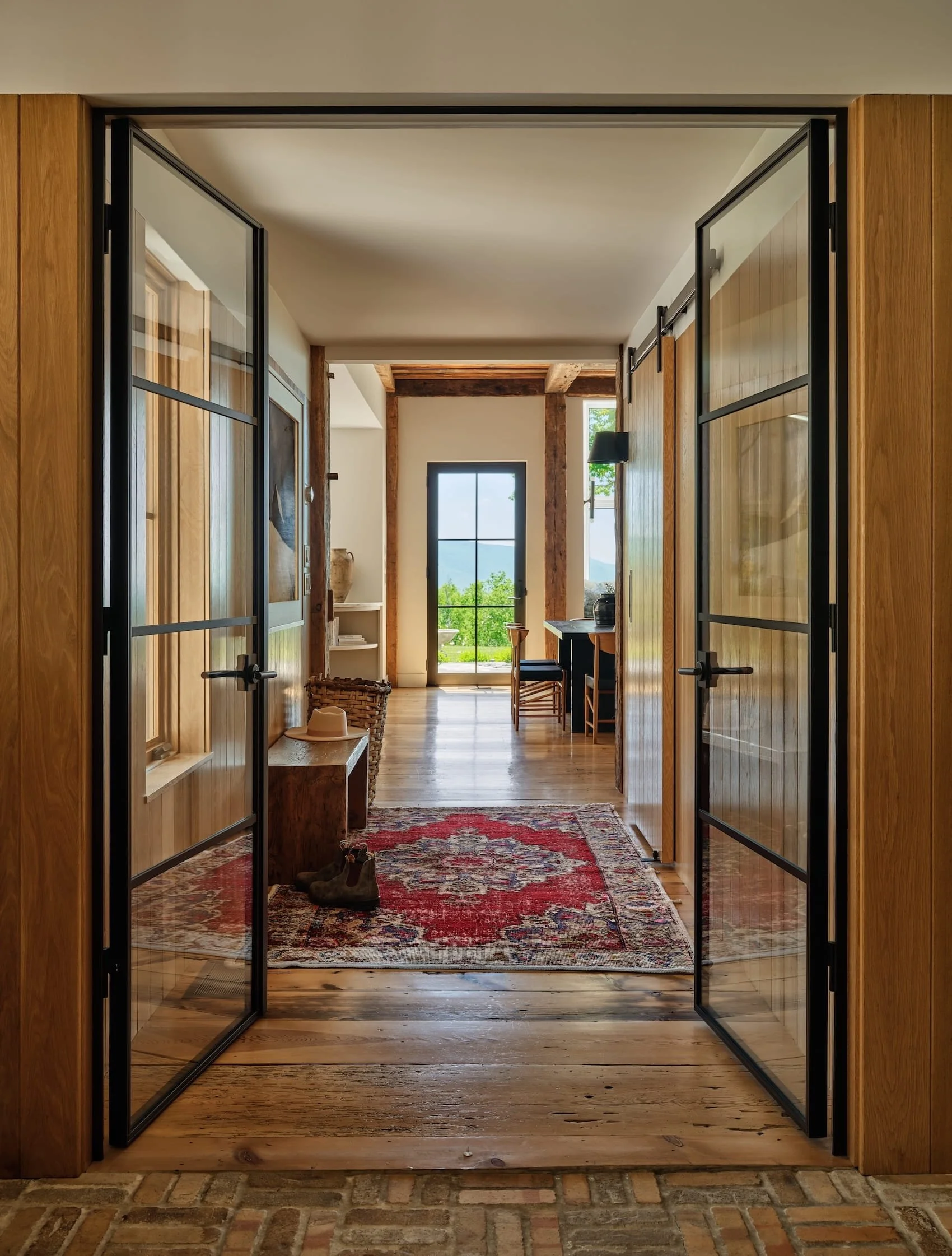 Hallway in a renovated home in Dorset, Vermont, with wood plank floors, dark metal-framed glass doors, and a red patterned runner leading to a light-filled room beyond.