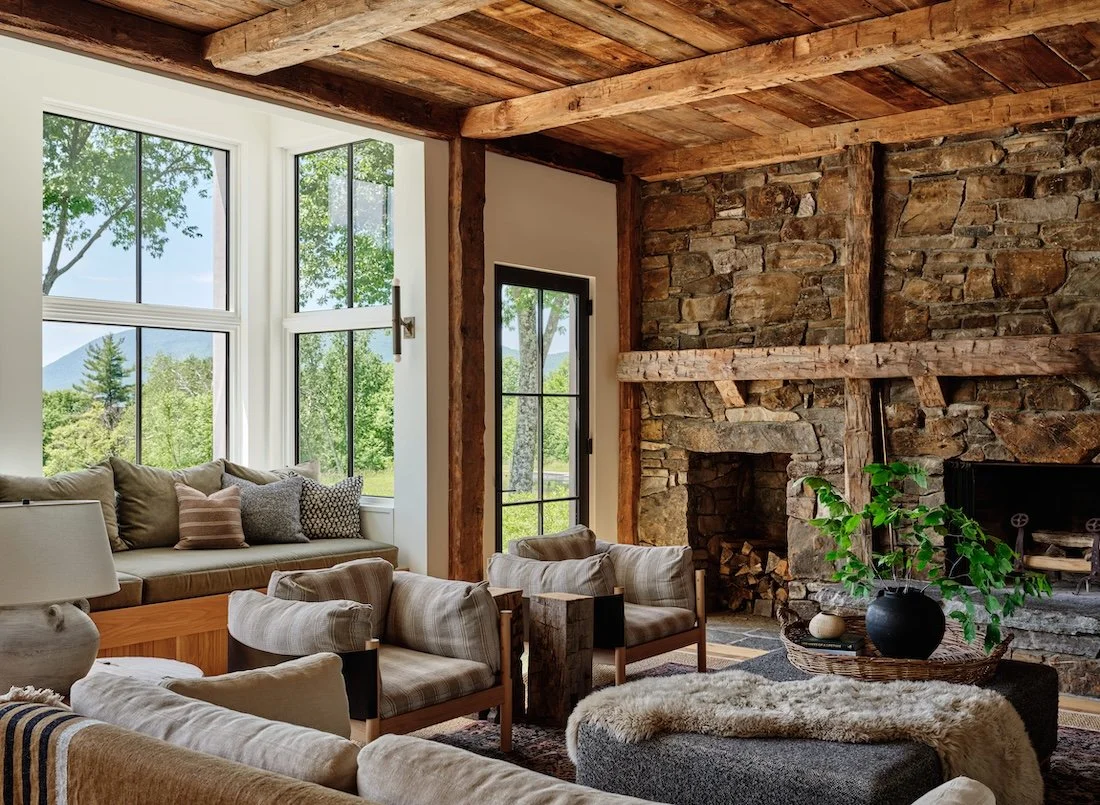 Living room in a renovated home in Dorset, Vermont, with a stone fireplace, exposed wood ceiling beams, large windows, and a seating area arranged around the hearth.