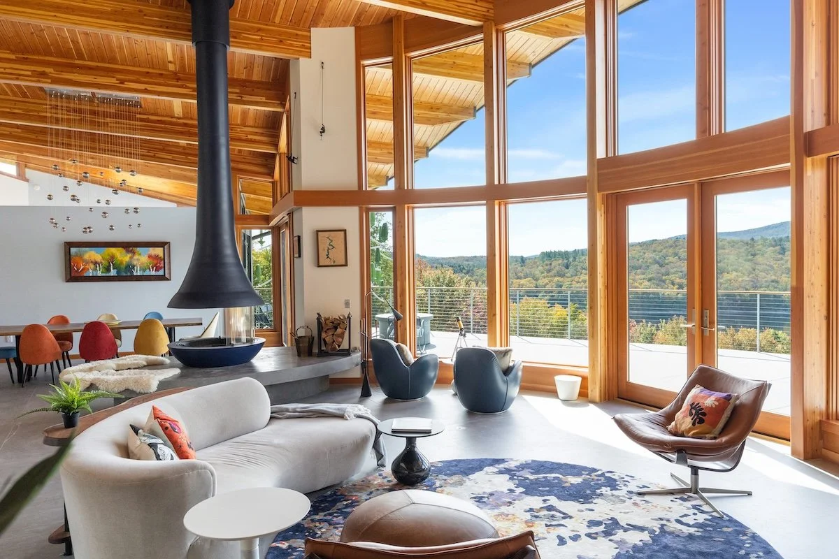 Living room in a custom home in Wilmington, Vermont, with curved seating, a suspended fireplace, and tall wood-framed windows opening to lake and mountain views.